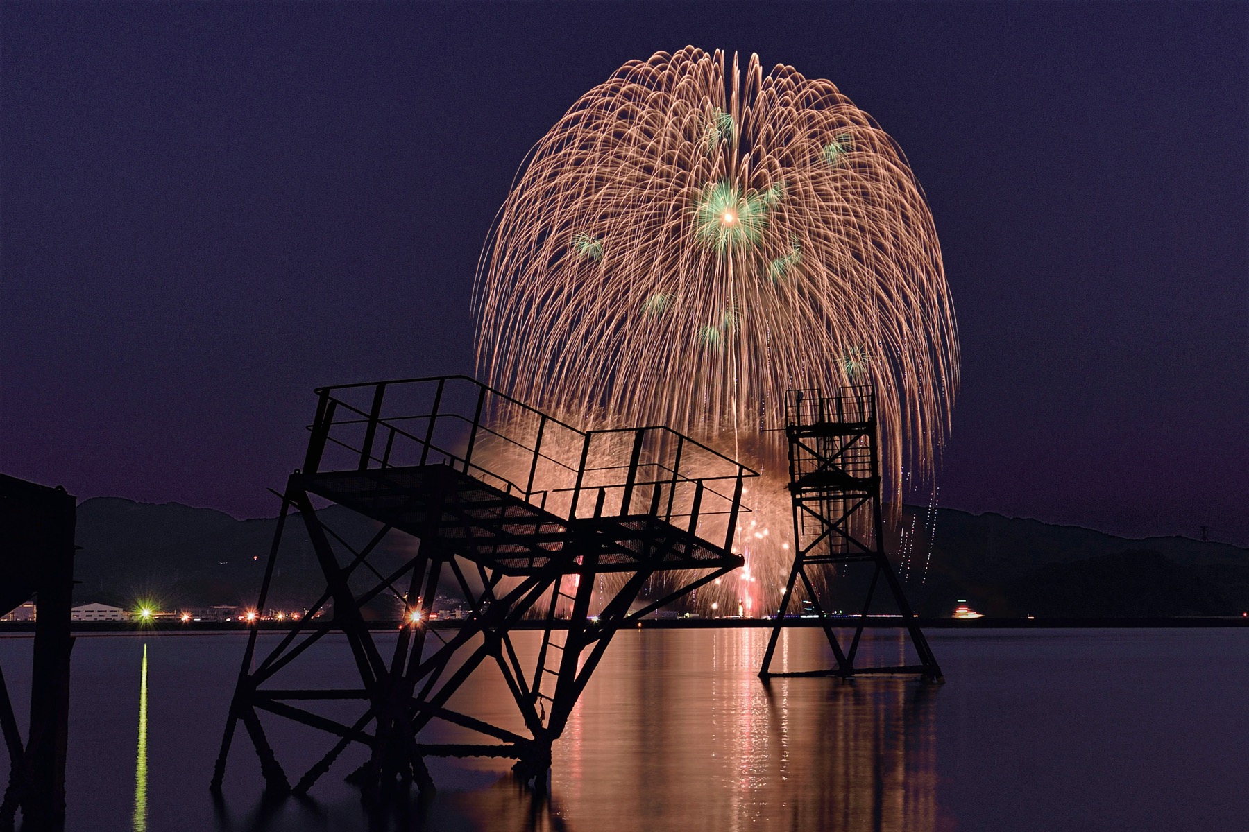 A large golden chrysanthemum firework bursting over Imari Bay at dusk behind the dark silhouette of a steel structure on the water, reflected in the calm bay at the Imari Bay fireworks festival in Saga, Japan