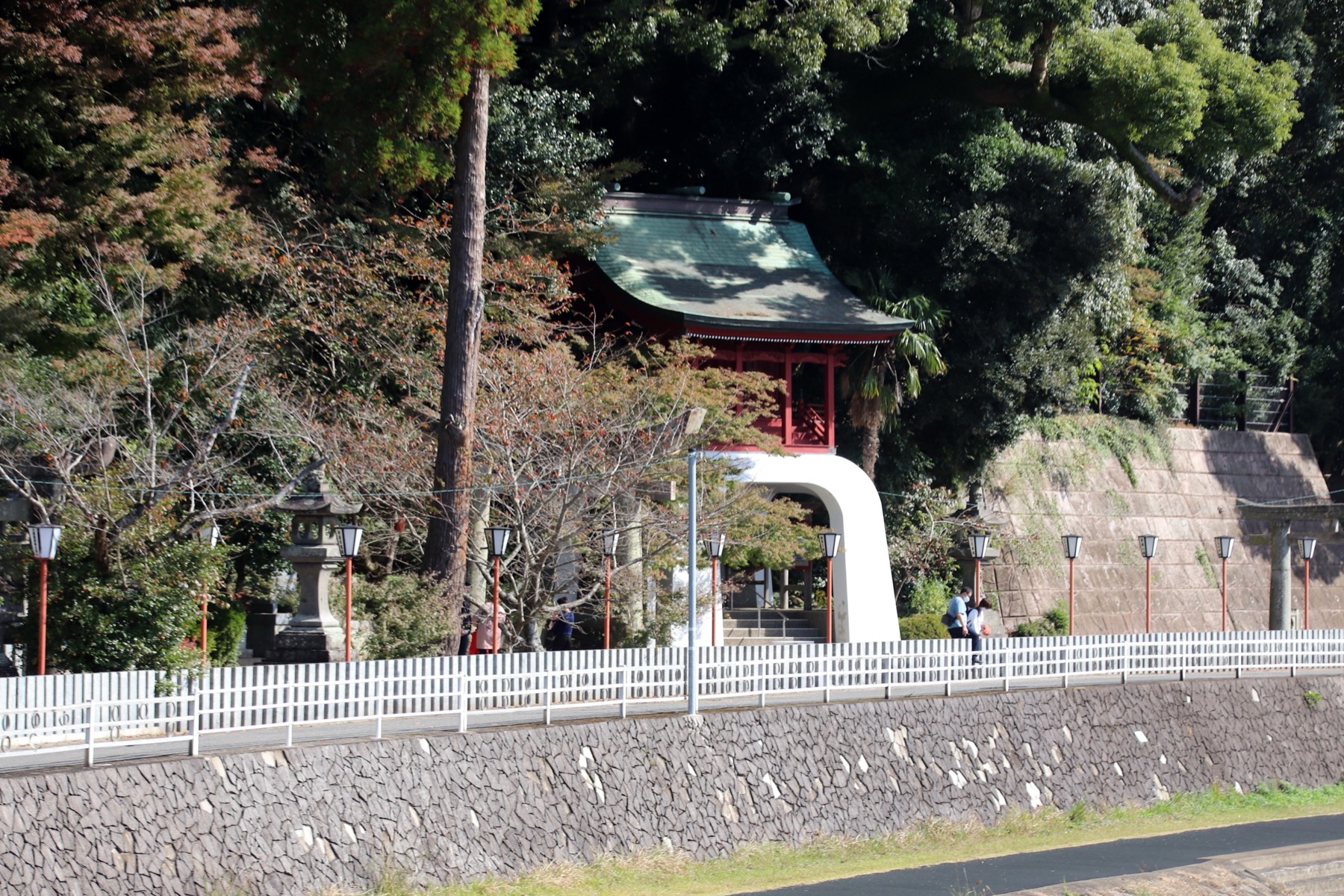 The vermilion tower gate of Imari Shrine seen across a stone-embanked river lined with red lanterns and autumn trees in Imari, Saga