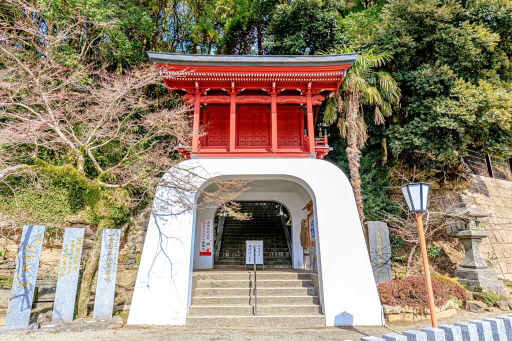 The striking vermilion two-storey tower gate (romon) of Imari Shrine in Imari, Saga, built over a white arched tunnel with stone steps leading up to the shrine, framed by trees