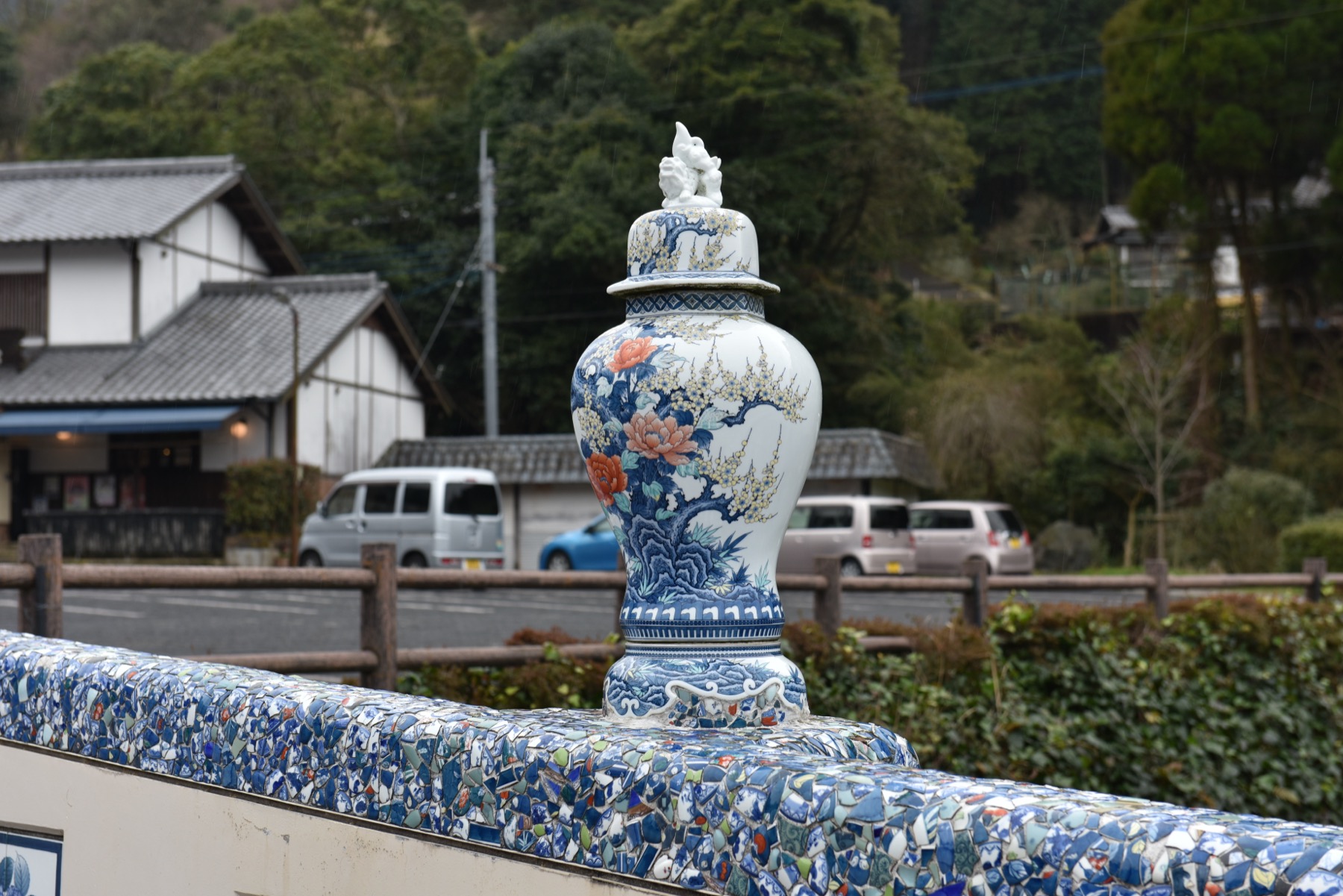 A large blue-and-white porcelain vase with a lion-dog lid displayed on a railing decorated with porcelain shards in a kiln village of the Imari area of Saga, houses and wooded hills behind