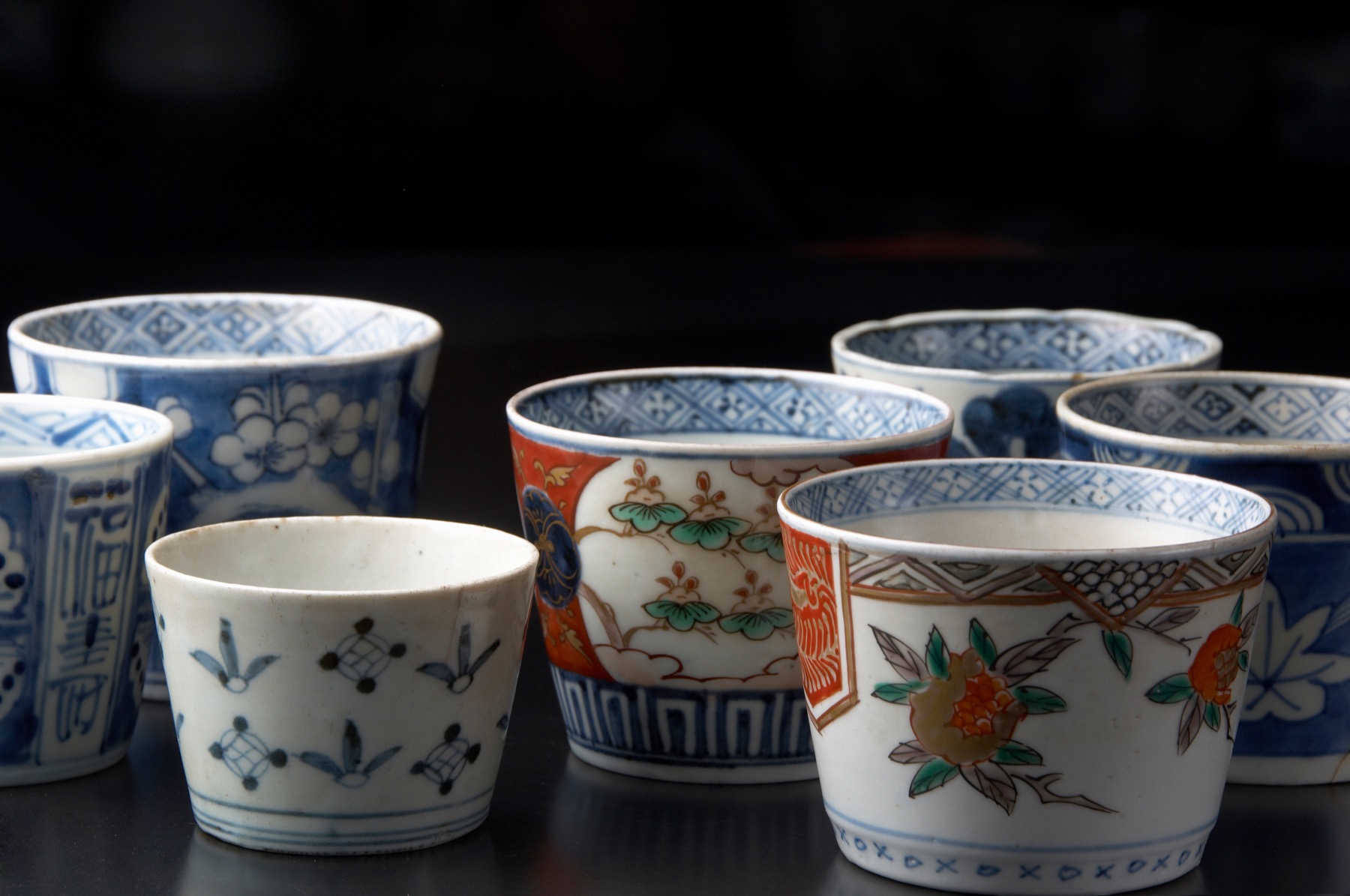 A collection of antique Imari soba-choko porcelain cups on a dark background, blue-and-white sometsuke and red-green overglaze patterns of flowers and geometric borders, made in the Arita and Imari area of Saga
