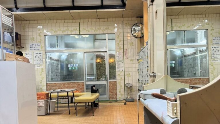 The interior of a traditional Japanese sento public bathhouse seen from the changing room, with tiled walls, mirrors, a glass partition marked open-air bath, a low yellow bench and a convex mirror.