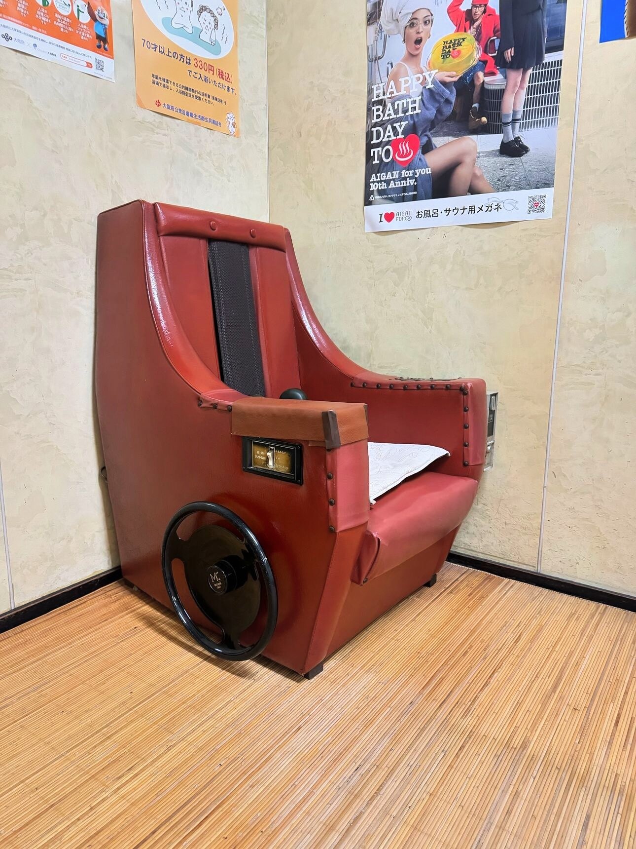 A retro red coin-operated reclining massage chair in the lounge corner of a Japanese sentō, beside a Happy Bath Day poster.