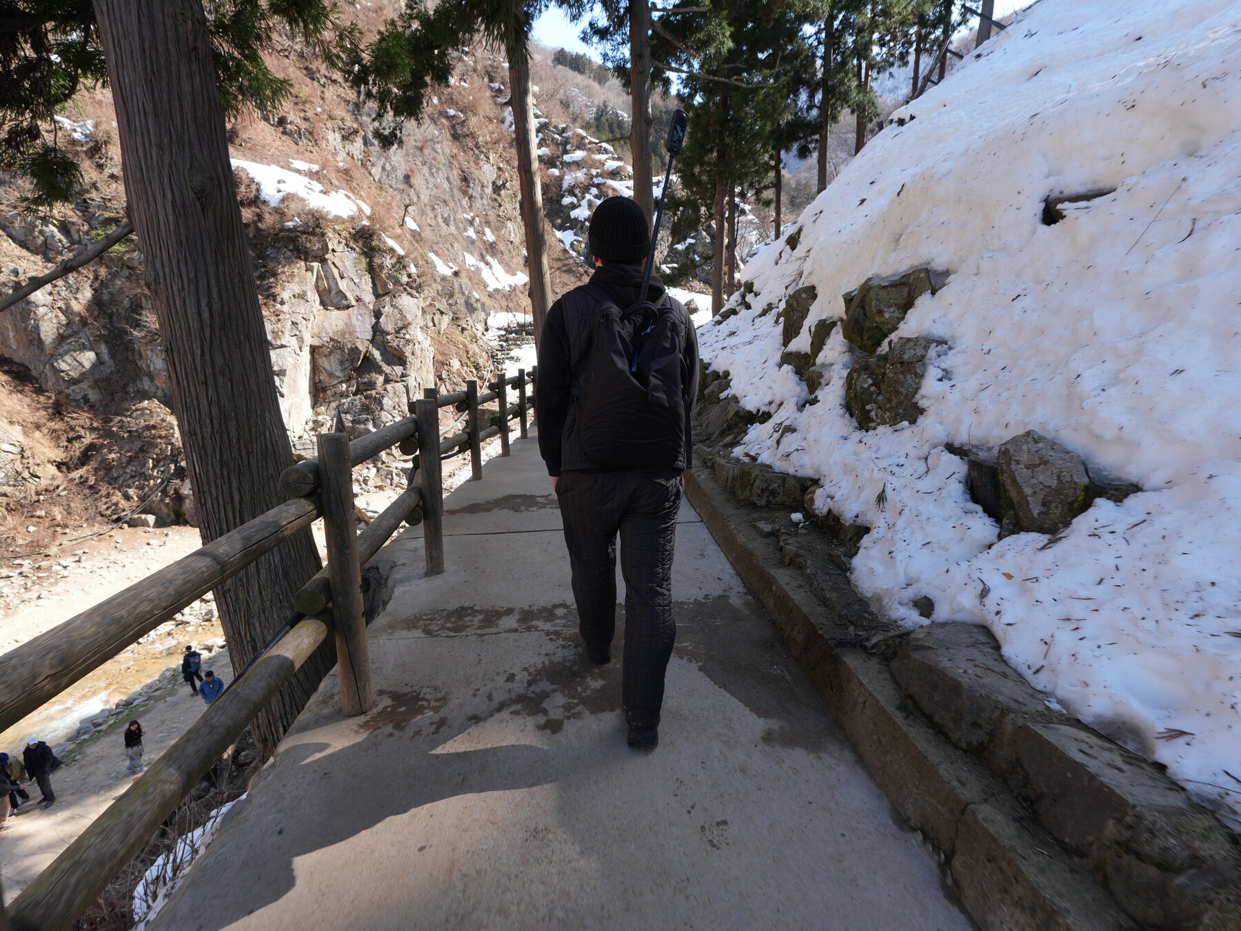 A visitor in winter clothing walking the icy, snow-packed forest trail to Jigokudani Snow Monkey Park, with a wooden railing on one side and a steep snowy bank on the other.