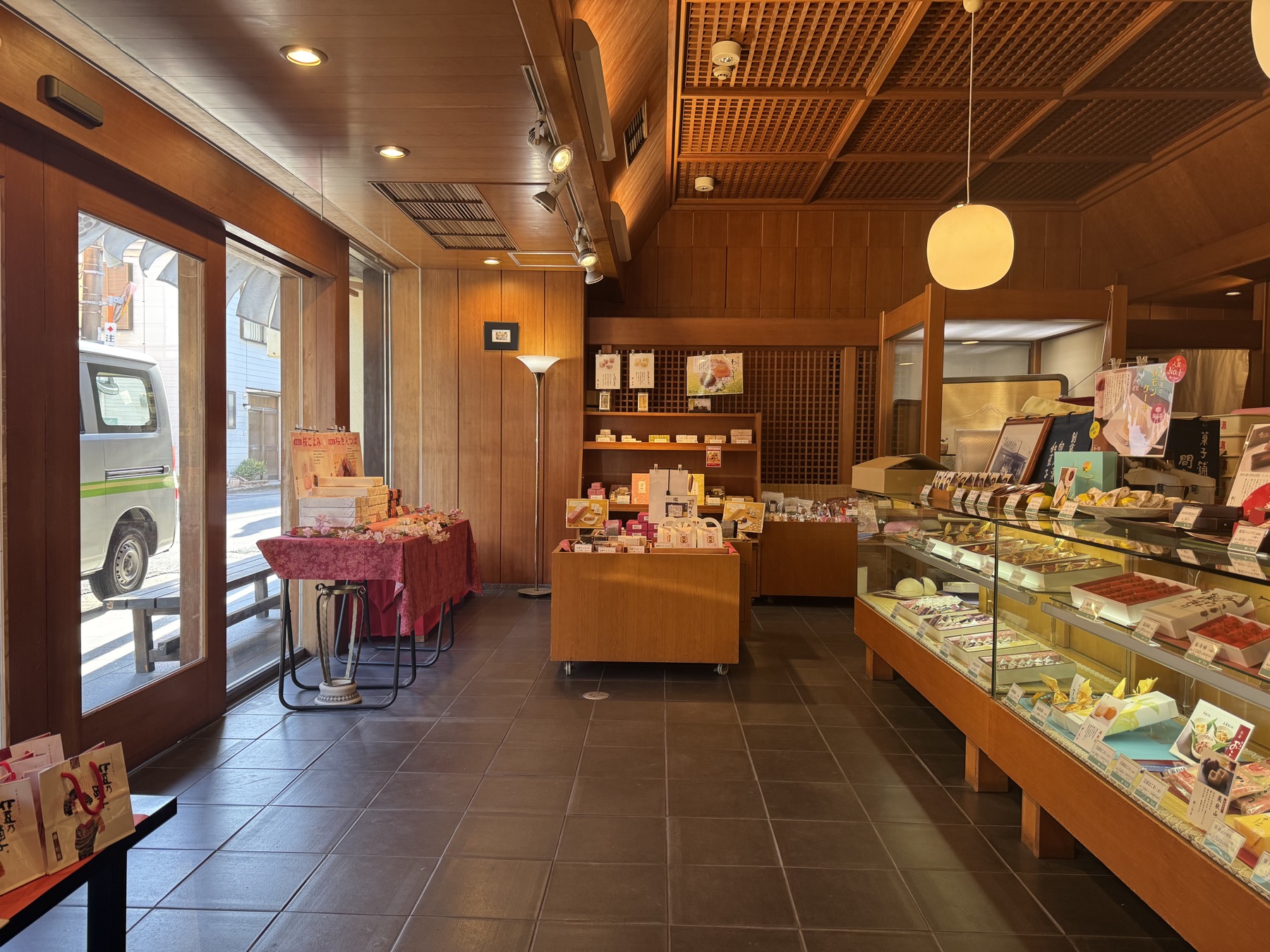 The warm wood-panelled interior of Kashiho Mase wagashi shop in Izu, Shizuoka, with glass display cases of Japanese sweets and a red-clothed product table by the window.