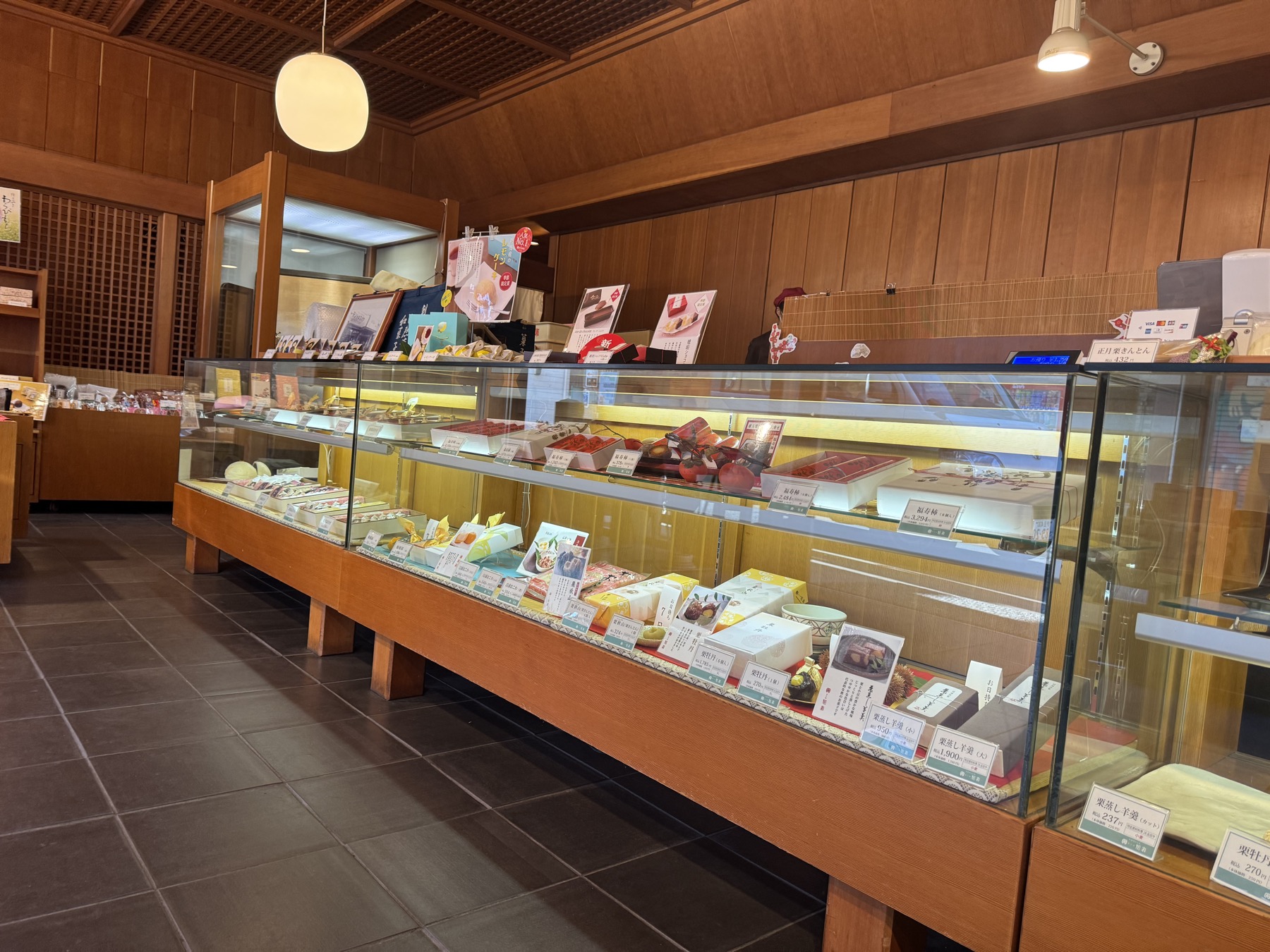 Glass display cases of seasonal Japanese sweets with handwritten price cards along the counter of Kashiho Mase wagashi shop in Izu, Shizuoka.