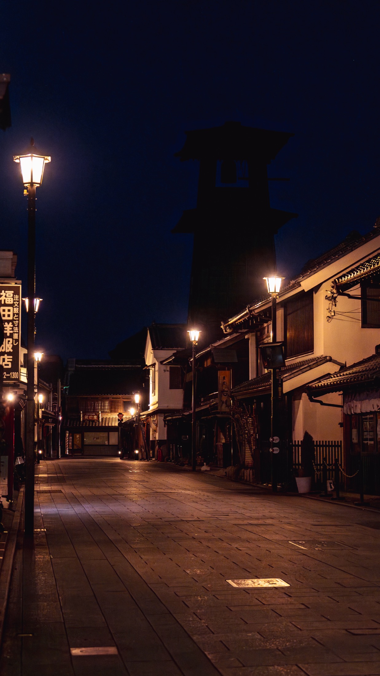 Kawagoe's Kurazukuri warehouse street at blue hour, completely empty, lit by street lamps with the dark silhouette of the Toki no Kane bell tower rising at the far end under a deep blue sky.
