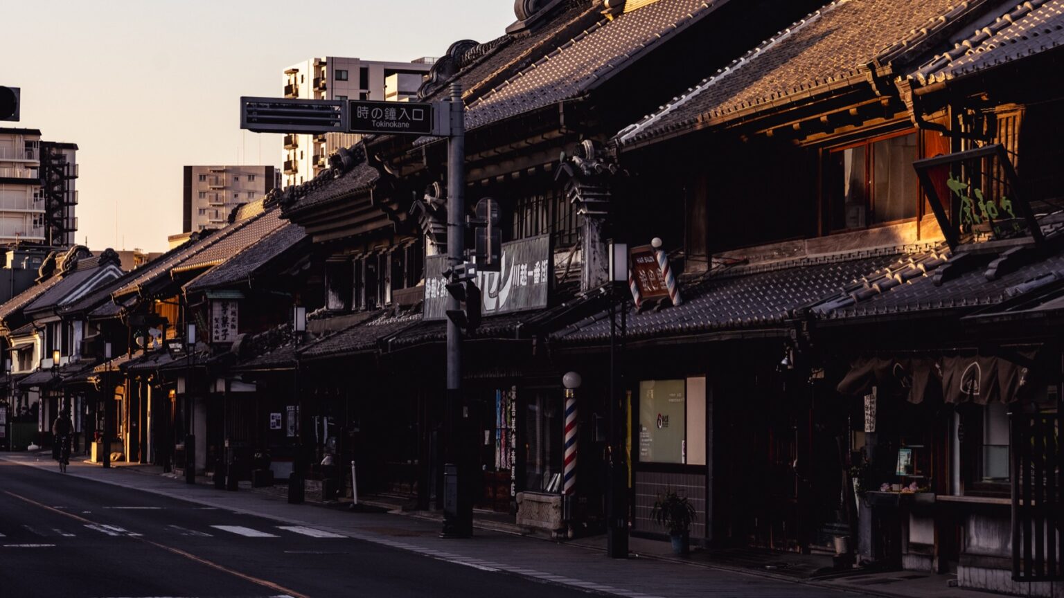 A row of black kurazukuri warehouses in Kawagoe lit by warm golden-hour light, with a small striped barber pole and old signage along an almost empty road.