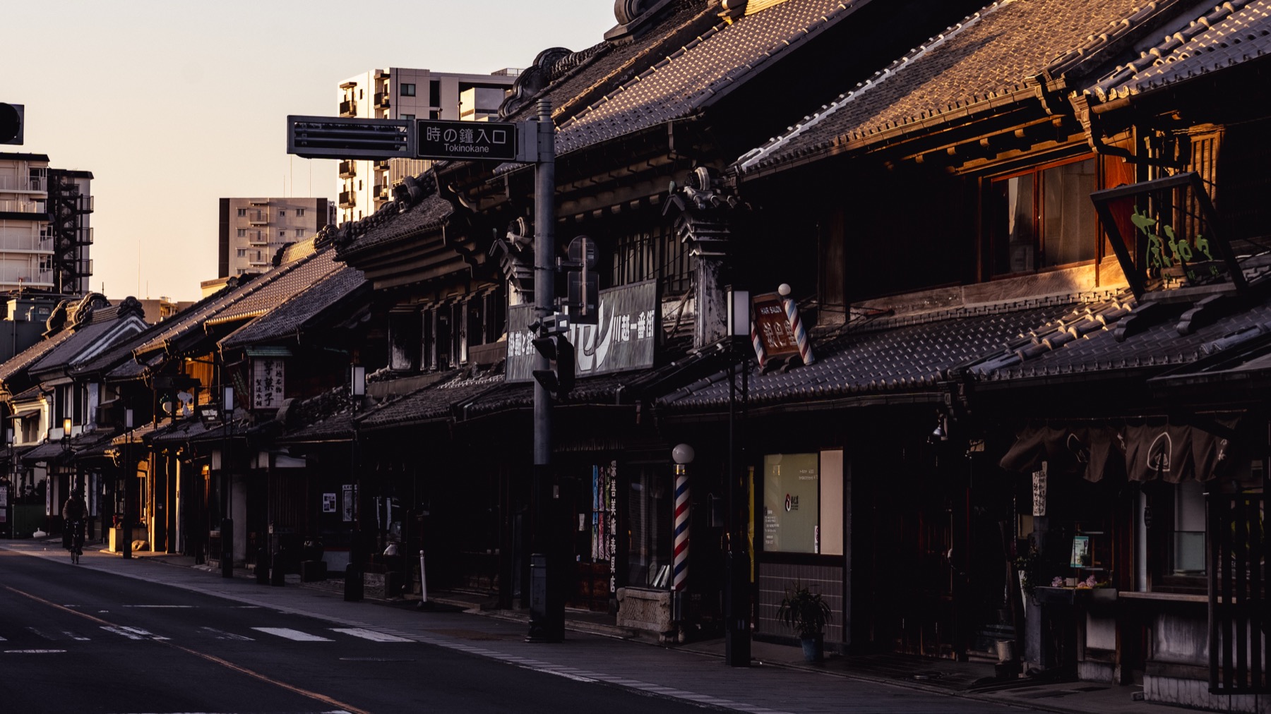 A row of black kurazukuri warehouses in Kawagoe lit by warm golden-hour light, with a small striped barber pole and old signage along an almost empty road.