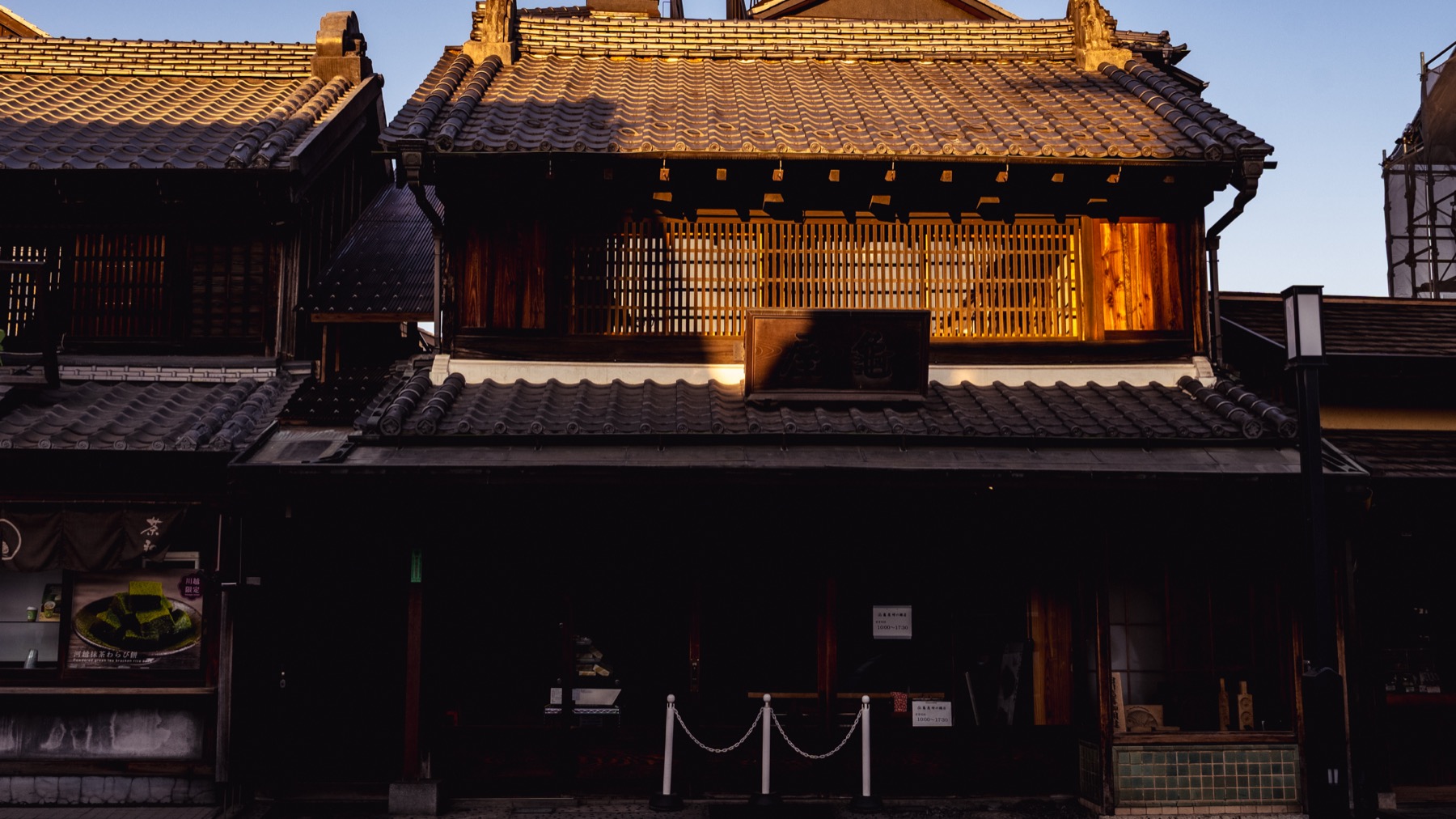 Close view of a Kawagoe kurazukuri warehouse facade in golden afternoon light, showing thick black tiled eaves, a timber lattice front and white-edged plaster walls.