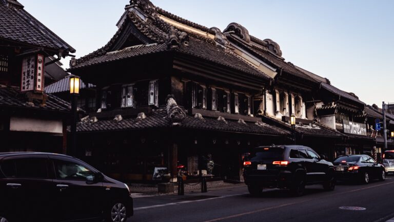 A large black clay-walled kurazukuri merchant warehouse on Ichibangai street in Kawagoe, with a heavy tiled roof and ornate gables, cars passing on the road in front under a clear sky.