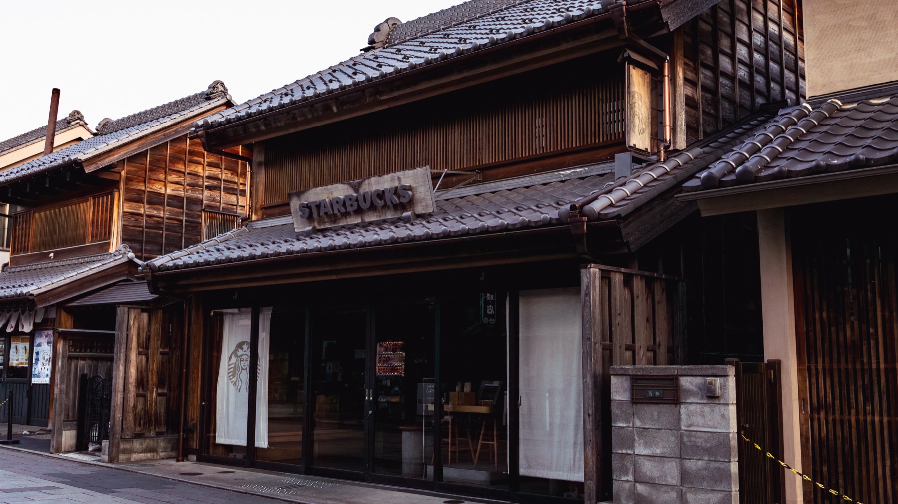 A timber-fronted kurazukuri storefront in Kawagoe in soft morning light, with a tiled roof, noren curtain and lattice windows, no people around.