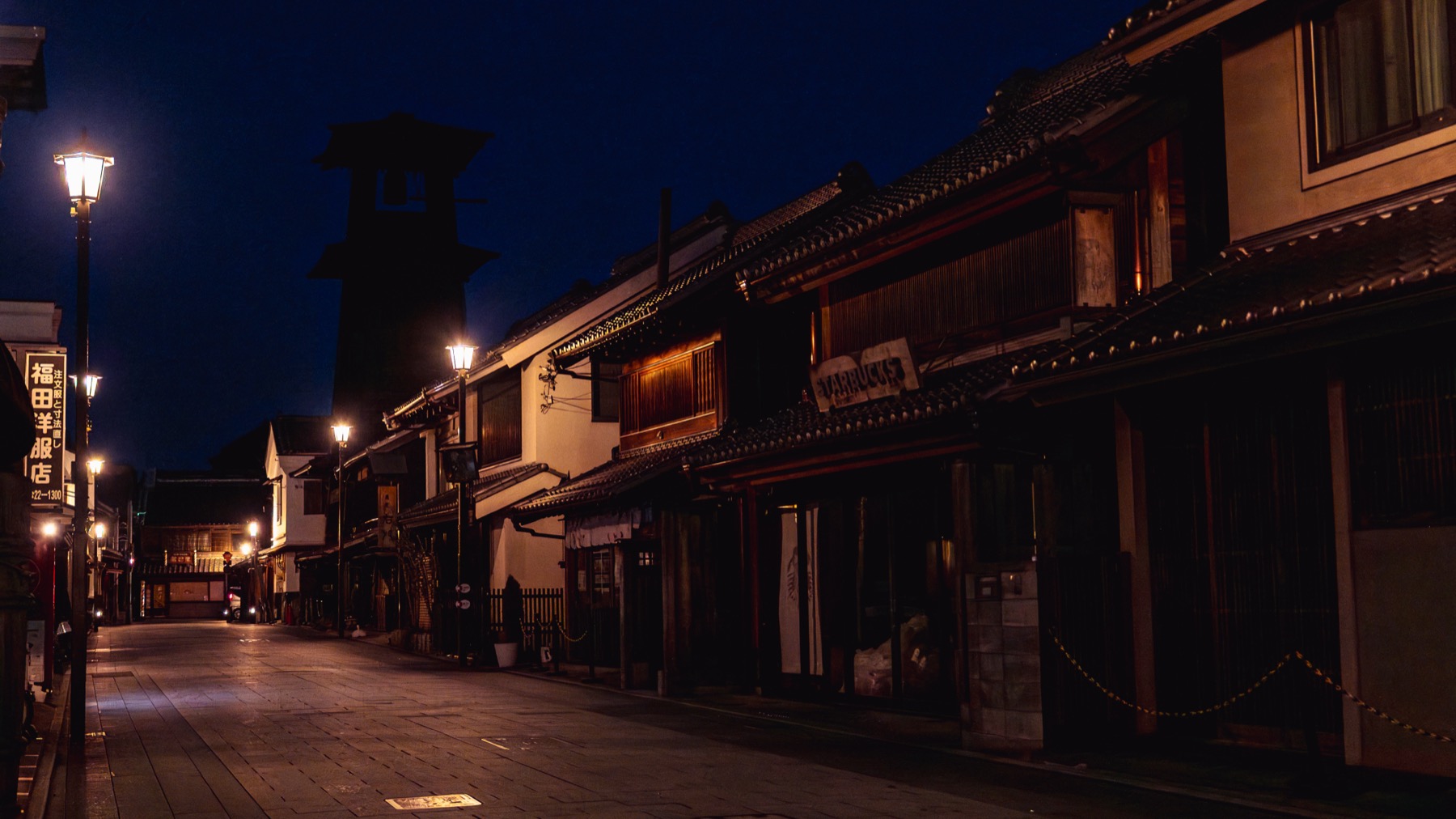 Ichibangai street in Kawagoe at blue hour, the warehouse street empty and lamplit with the Toki no Kane bell tower glowing softly at the far end under a deep blue sky.
