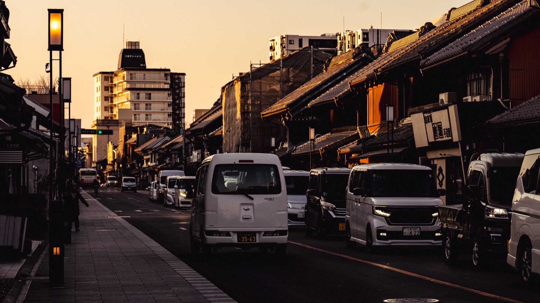 Ichibangai street in Kawagoe at golden hour, black kurazukuri warehouses lining the right side with street lamps, cars and a hotel building catching warm low sunlight.