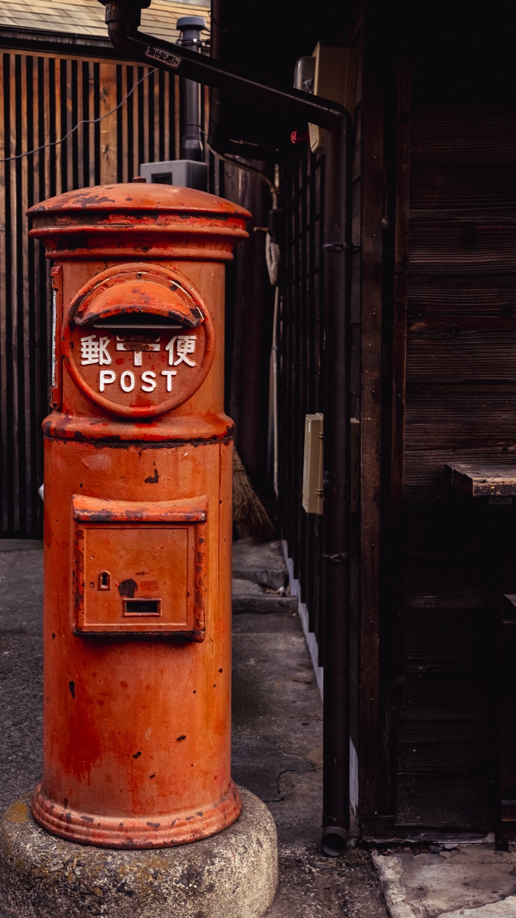 An old cylindrical red Japanese postbox marked POST standing on the cobbled street beside a dark kurazukuri building in Kawagoe.