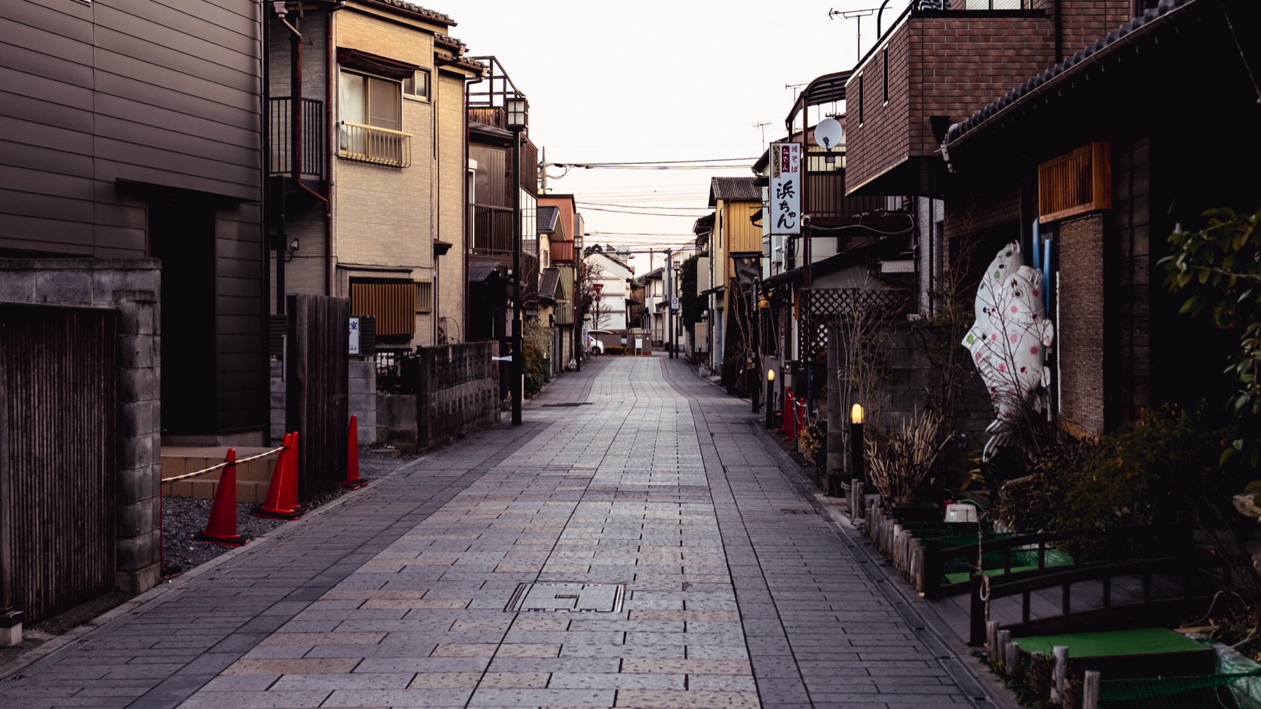 A quiet stone-paved side street in Kawagoe lined with mixed old buildings, traffic cones and a brick building, almost no people, soft overcast light.