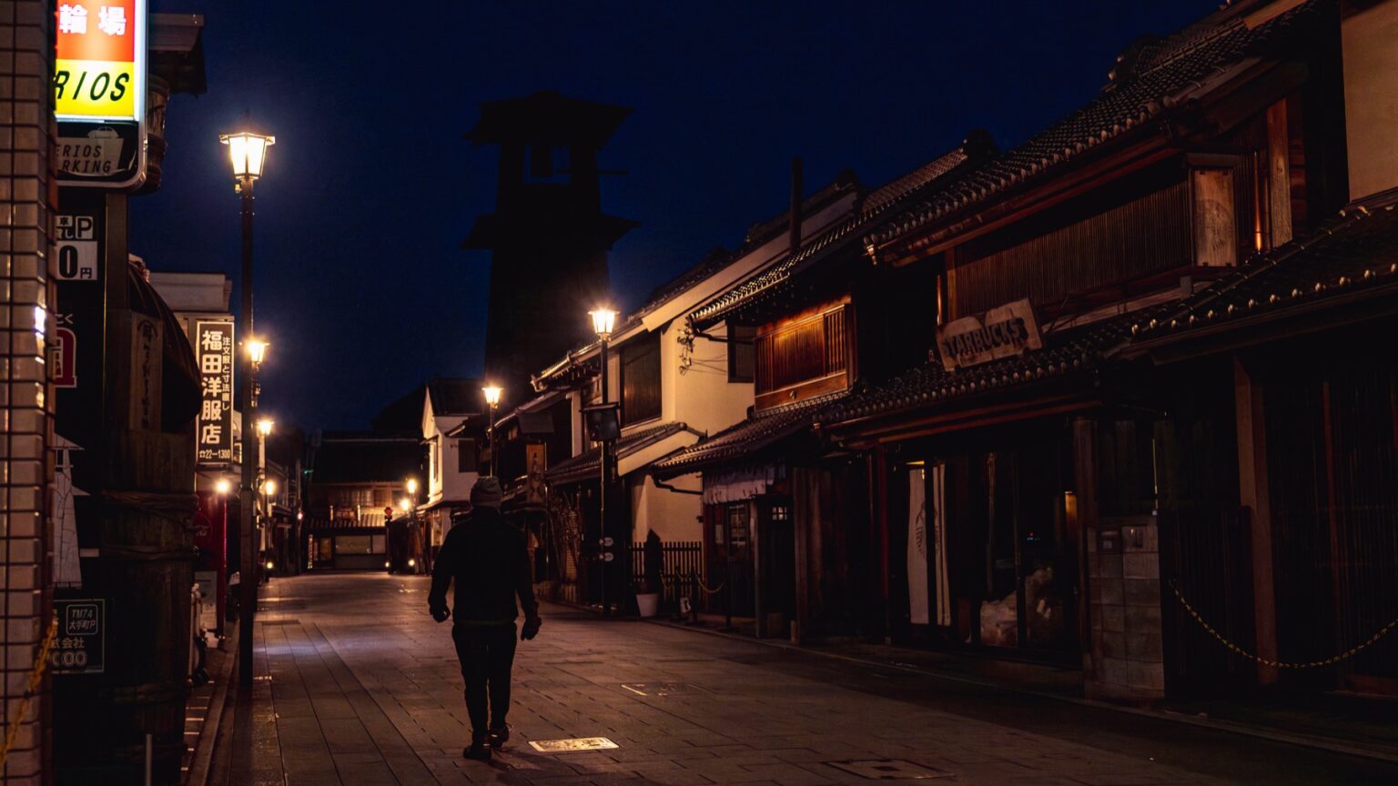 The Toki no Kane bell tower at blue hour in Kawagoe, a lone person walking down the lamplit empty warehouse street toward the dark tower under a deep blue night sky.