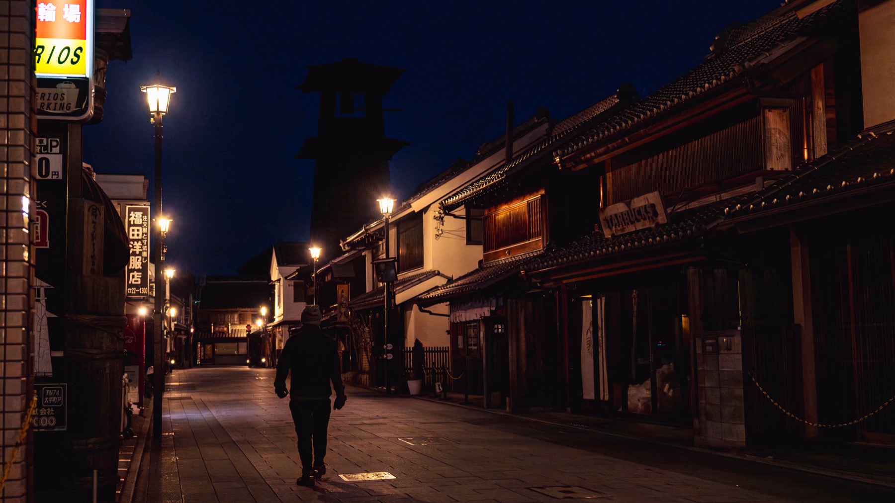 The Toki no Kane bell tower at blue hour in Kawagoe, a lone person walking down the lamplit empty warehouse street toward the dark tower under a deep blue night sky.