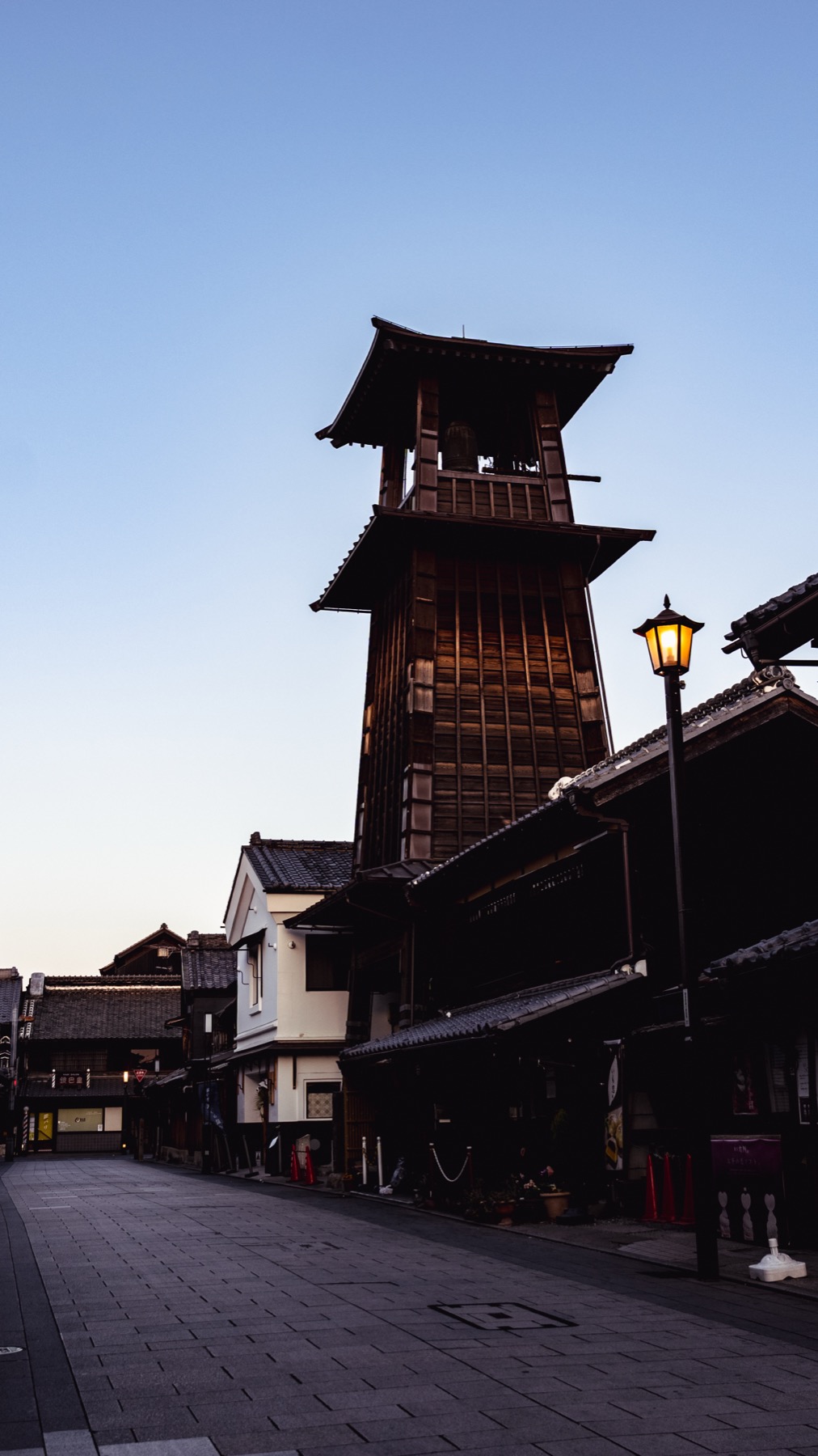 The Toki no Kane bell tower at dawn in Kawagoe, the wooden tower against a soft pale-blue sky with an empty cobbled street and a lit street lamp below.
