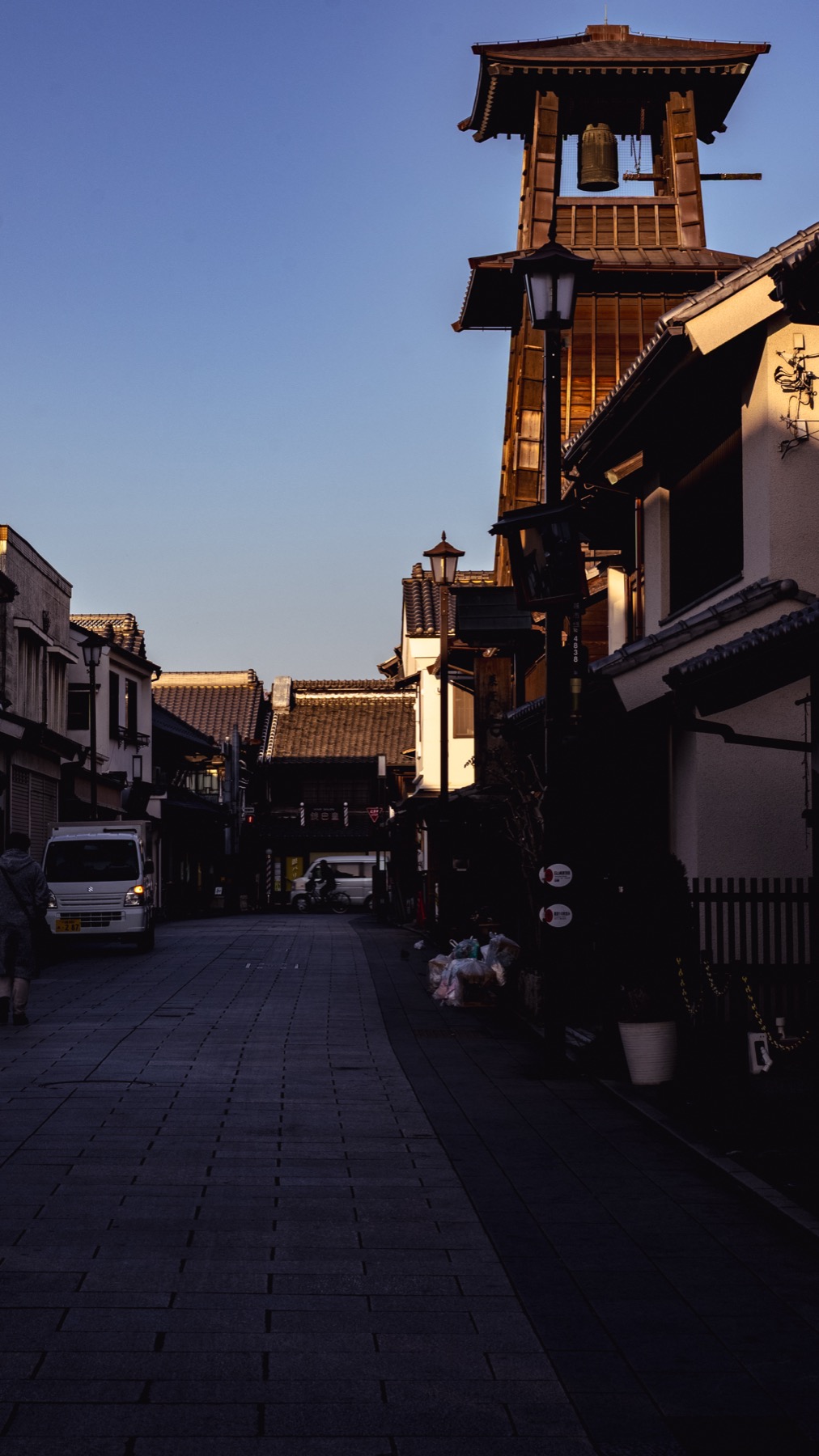 The Toki no Kane bell tower seen from the cobbled Kurazukuri street in Kawagoe, warm afternoon light on the timber, an old street lamp and white-walled storehouses alongside.