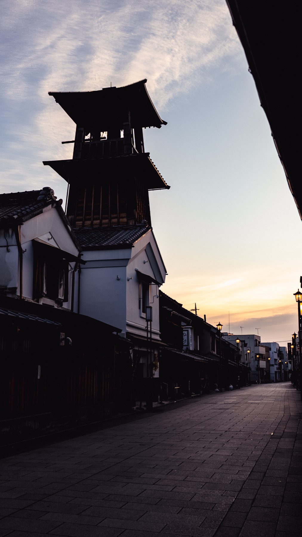 The Toki no Kane bell tower silhouetted black against an orange and blue sunset sky in Kawagoe, looking down an empty street lined with white-walled storehouses.
