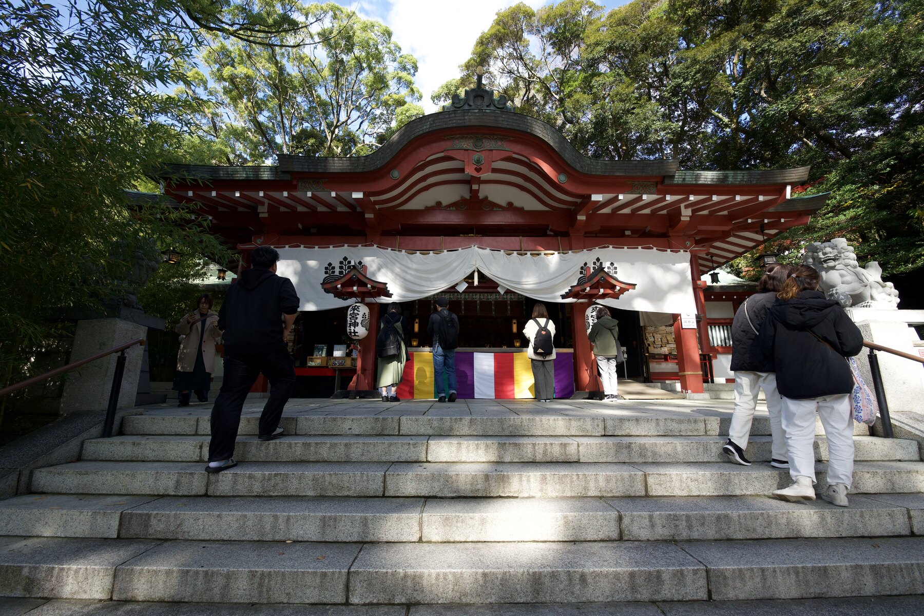 The vermilion main hall of Kinomiya Shrine in Atami at the top of a flight of stone steps, with a white curtain across the front and visitors approaching to pray.