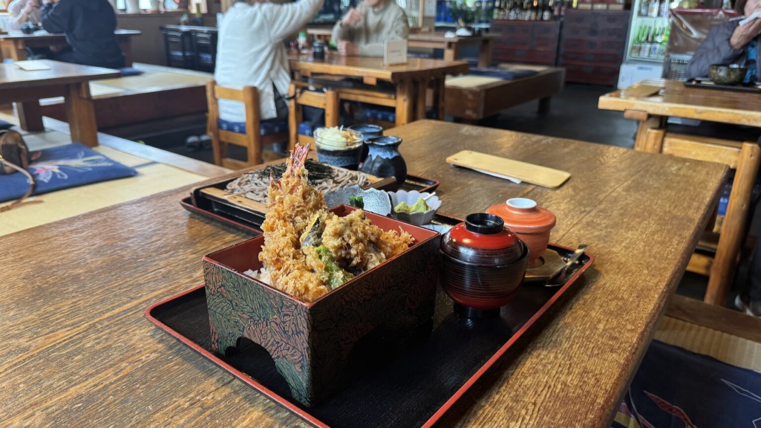 The tempura-rice box and a basket of hand-cut soba on a wooden table in the rustic dining room of Kintaro soba house in Gotemba, Shizuoka, with other diners and old beams behind