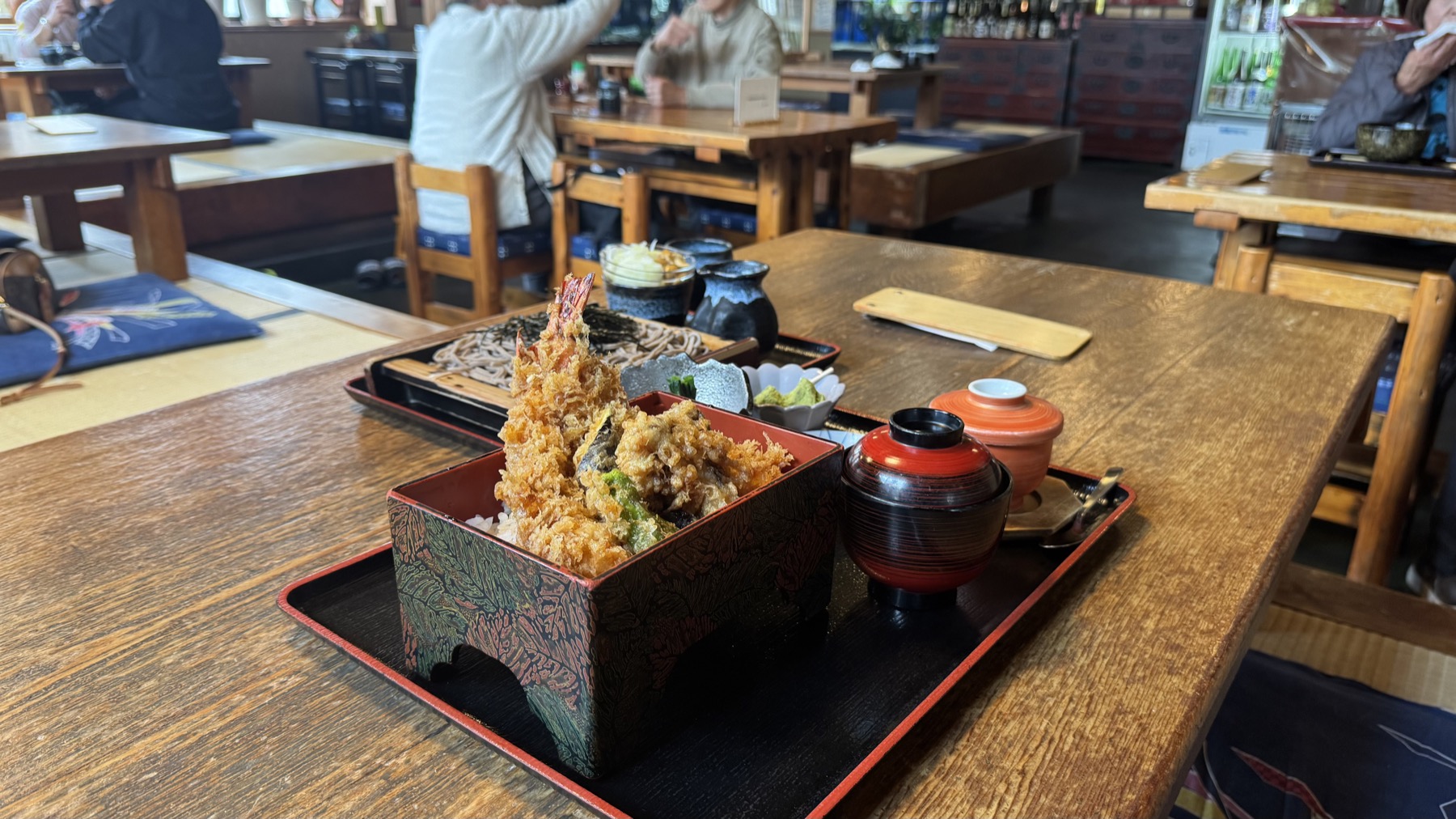 The tempura-rice box and a basket of hand-cut soba on a wooden table in the rustic dining room of Kintaro soba house in Gotemba, Shizuoka, with other diners and old beams behind