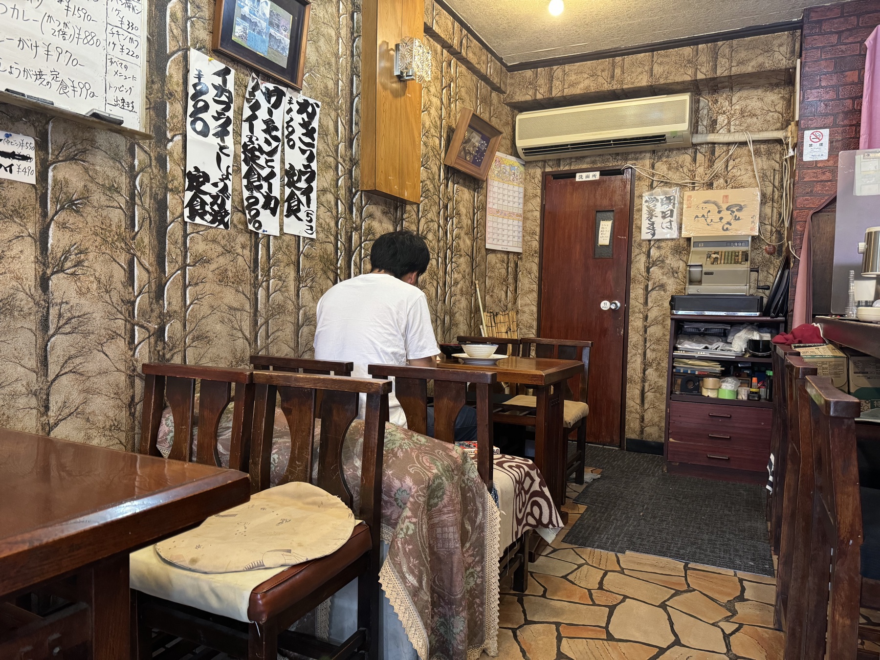 The retro interior of a small Tokyo diner: wood-grain walls hung with handwritten menu signs, a customer seated at a wooden table, a cash register, and a stone-tiled floor.