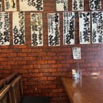 A brick-patterned wall inside the small Imahana diner in Setagaya, Tokyo, completely covered with hand-brushed paper menu cards listing set meals in black ink above a wooden table and bench.
