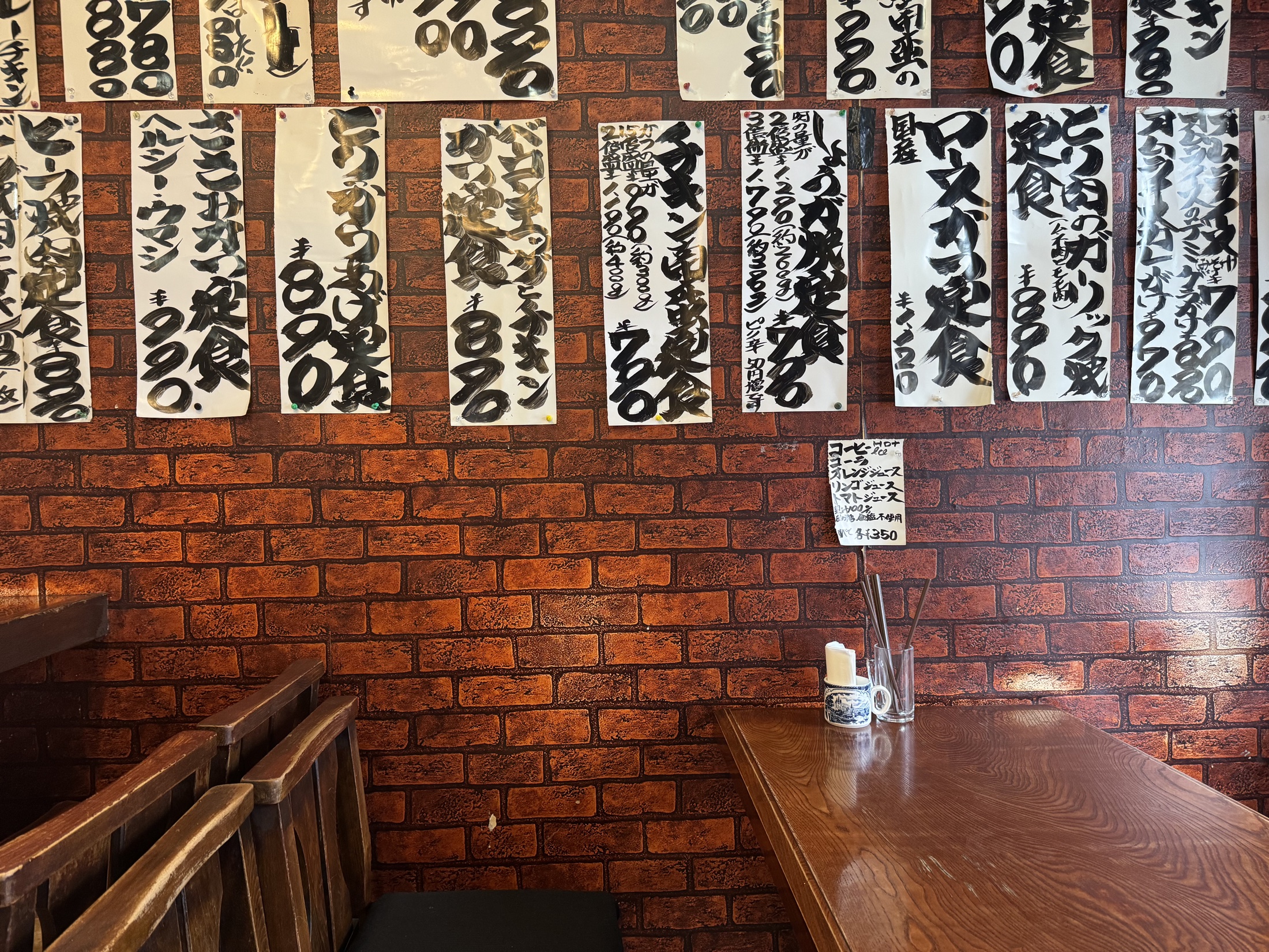 A brick-patterned wall inside the small Imahana diner in Setagaya, Tokyo, completely covered with hand-brushed paper menu cards listing set meals in black ink above a wooden table and bench.