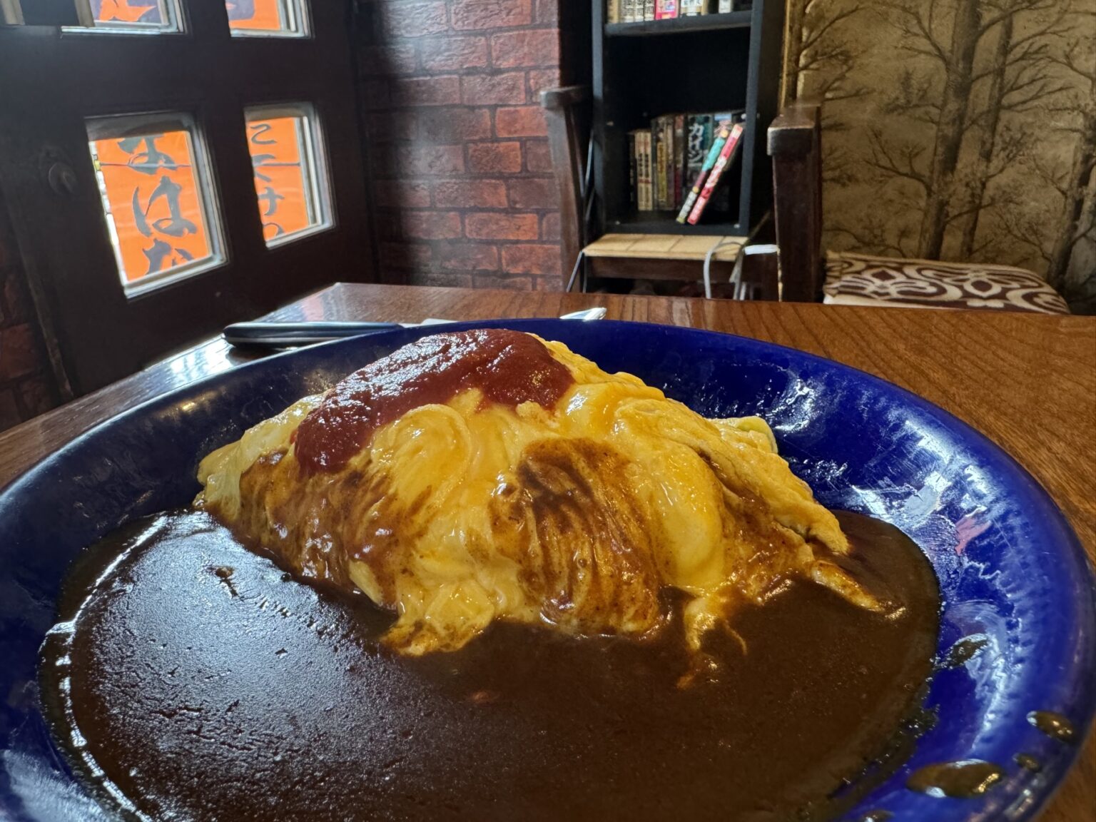 An omelette-wrapped rice omurice topped with a streak of ketchup, sitting in a pool of dark brown sauce on a deep blue plate, by a window etched with the shop name.