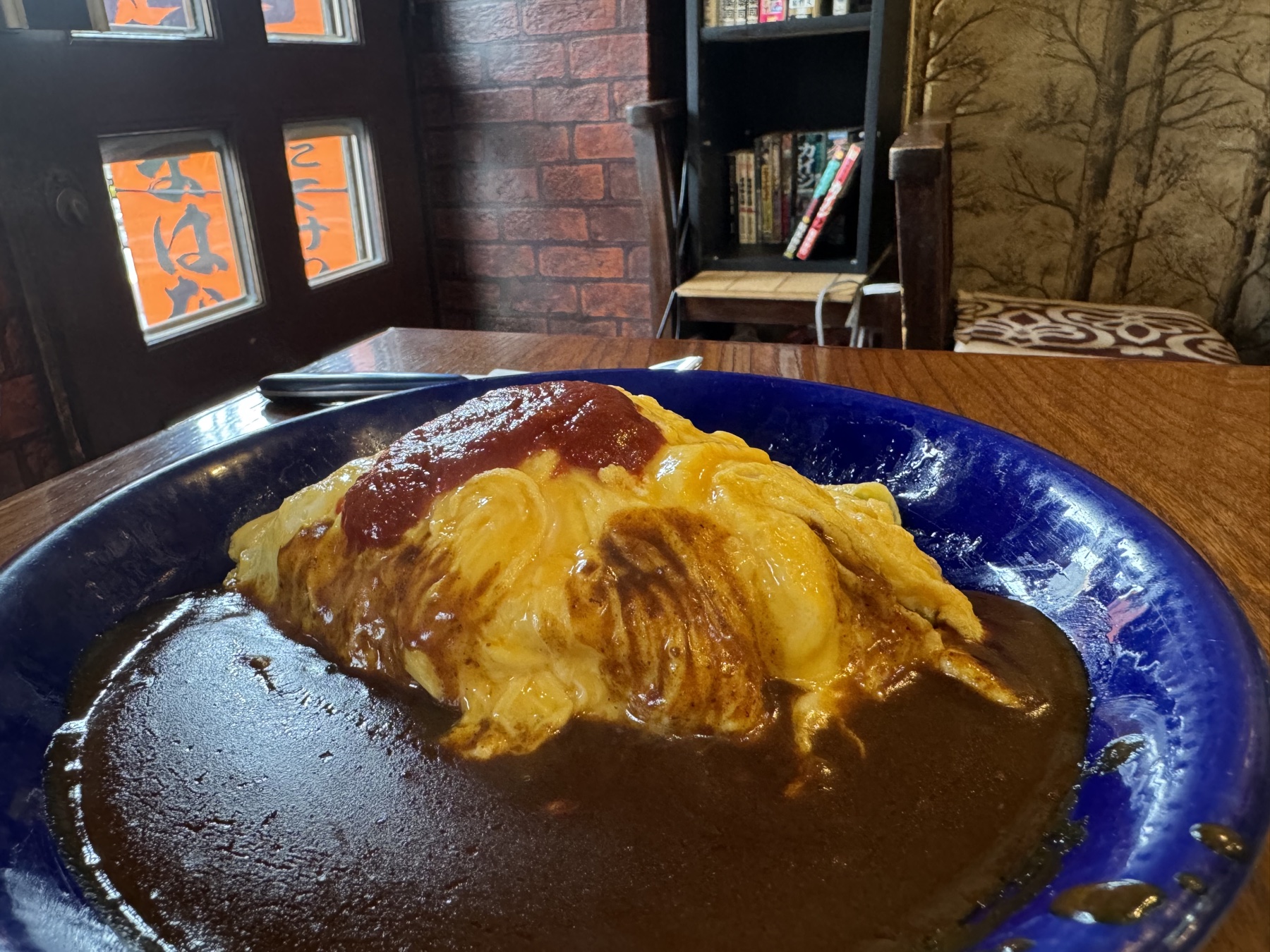 An omelette-wrapped rice omurice topped with a streak of ketchup, sitting in a pool of dark brown sauce on a deep blue plate, by a window etched with the shop name.