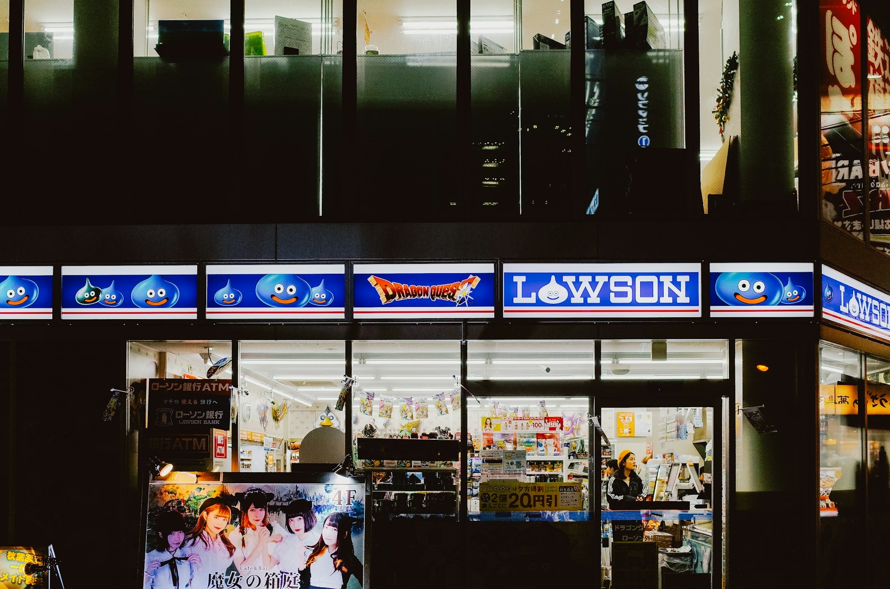A Lawson convenience store glowing blue at night in a Japanese city, the illuminated signage and glass storefront showing magazine racks, an in-store ATM and shelves of goods, open 24 hours