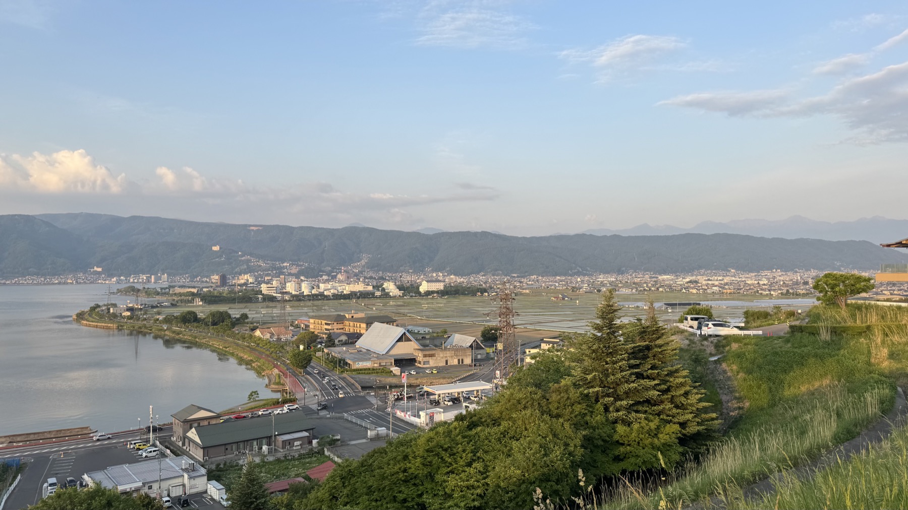 The eastern shore of Lake Suwa at dusk with rice fields and a lakeside road in the foreground and the town and mountains beyond under a fading sky.
