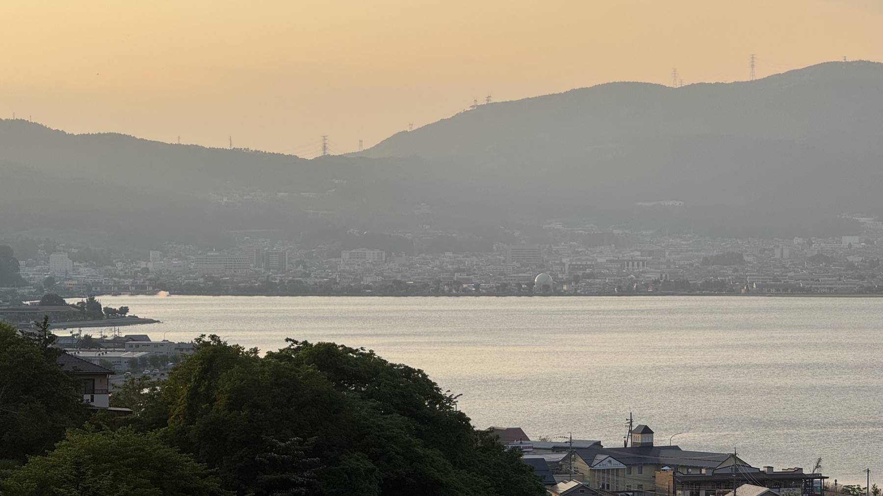 Lake Suwa glowing in golden-hour light with the town of Suwa spread along the far shore beneath hazy mountains, seen from a wooded hillside above the lake.