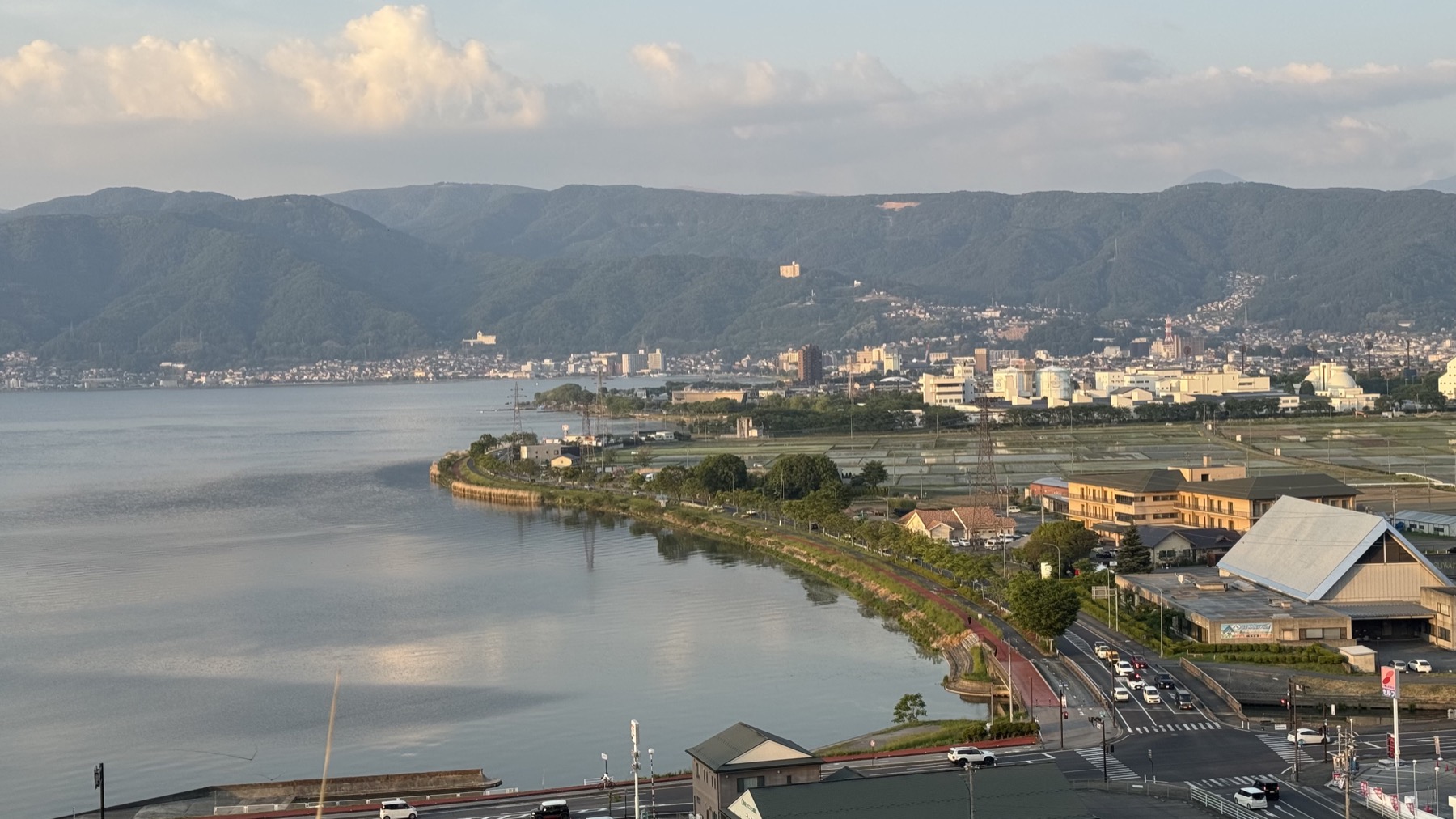The curving lakeshore of Lake Suwa with the flat 16 km cycling and jogging path running along the water, the town and mountains beyond, on a clear evening in Nagano.