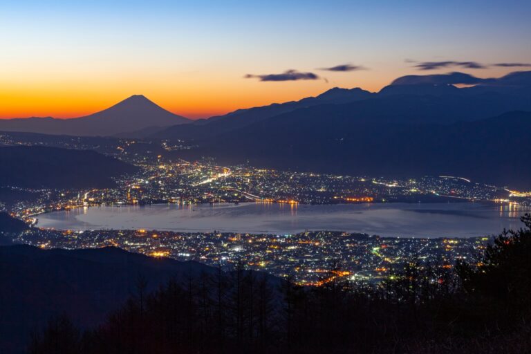 Mount Fuji silhouetted against an orange dusk sky beyond Lake Suwa, with the city lights of Suwa, Okaya and Shimosuwa ringing the dark water, seen from the hills above the lake in Nagano.