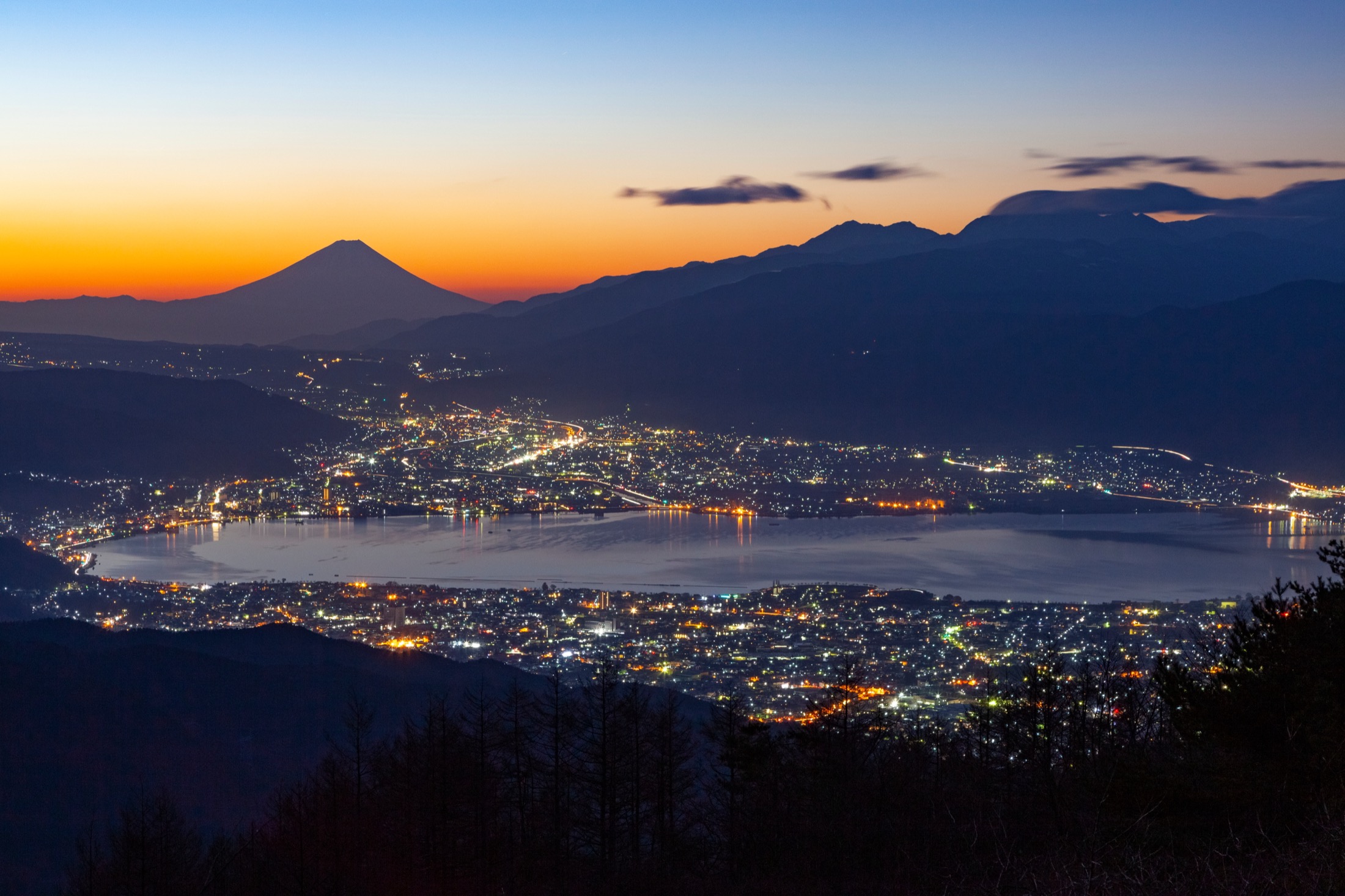 Mount Fuji silhouetted against an orange dusk sky beyond Lake Suwa, with the city lights of Suwa, Okaya and Shimosuwa ringing the dark water, seen from the hills above the lake in Nagano.