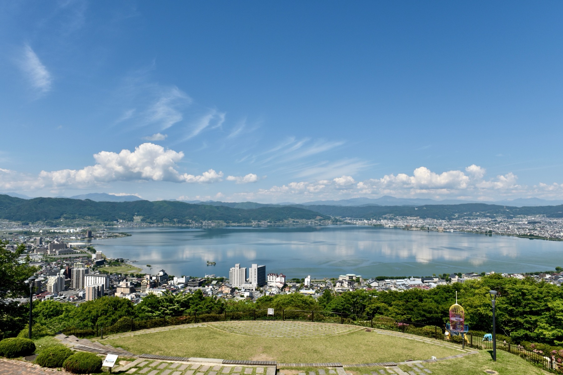 Lake Suwa seen on a clear summer day from Tateishi Park, a grassy hilltop terrace overlooking the lake and the town of Suwa, with mountains ringing the far shore in Nagano.