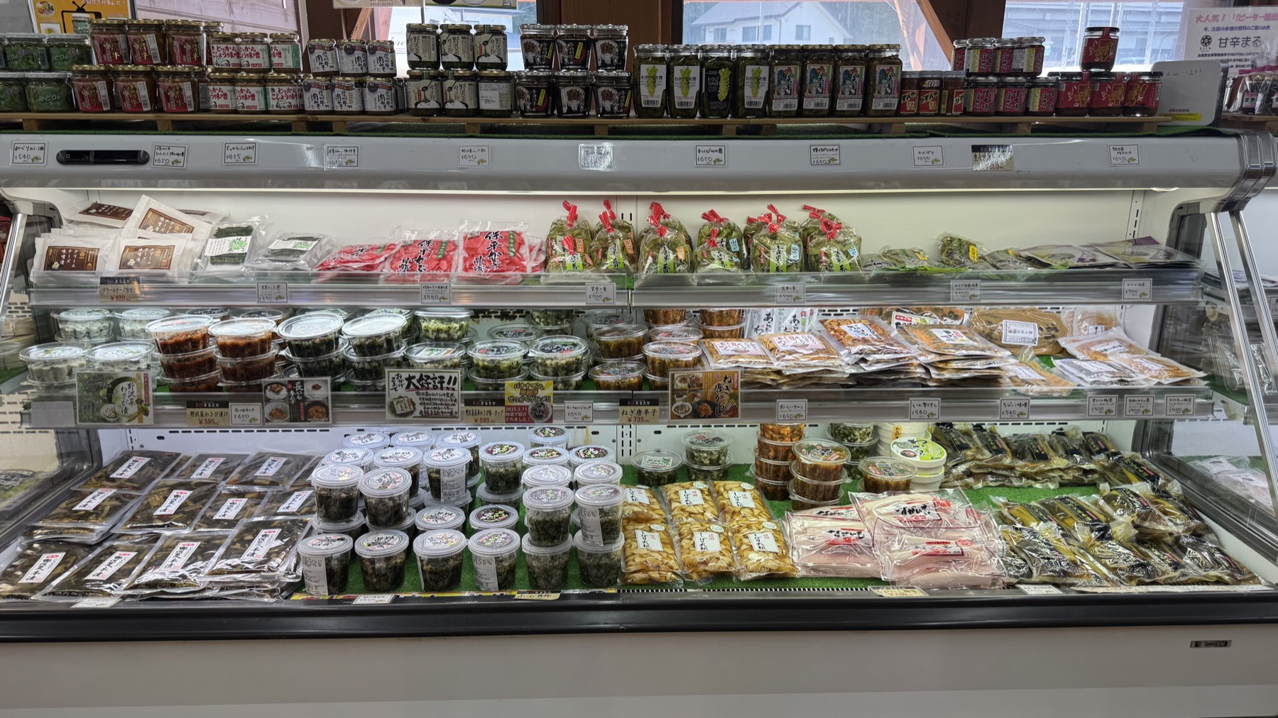 A refrigerated display case at Michi-no-Eki Hakuba filled with local prepared foods, including pickles, packaged vegetables, deli items and seasonal dishes arranged on several chilled shelves.