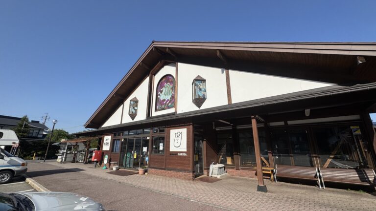 The chalet-style timber-and-plaster building of Michi-no-Eki Hakuba roadside station in Nagano under a clear blue sky, with stained-glass mountain artwork on the gable, a HAKUBA sign and a car park out front.