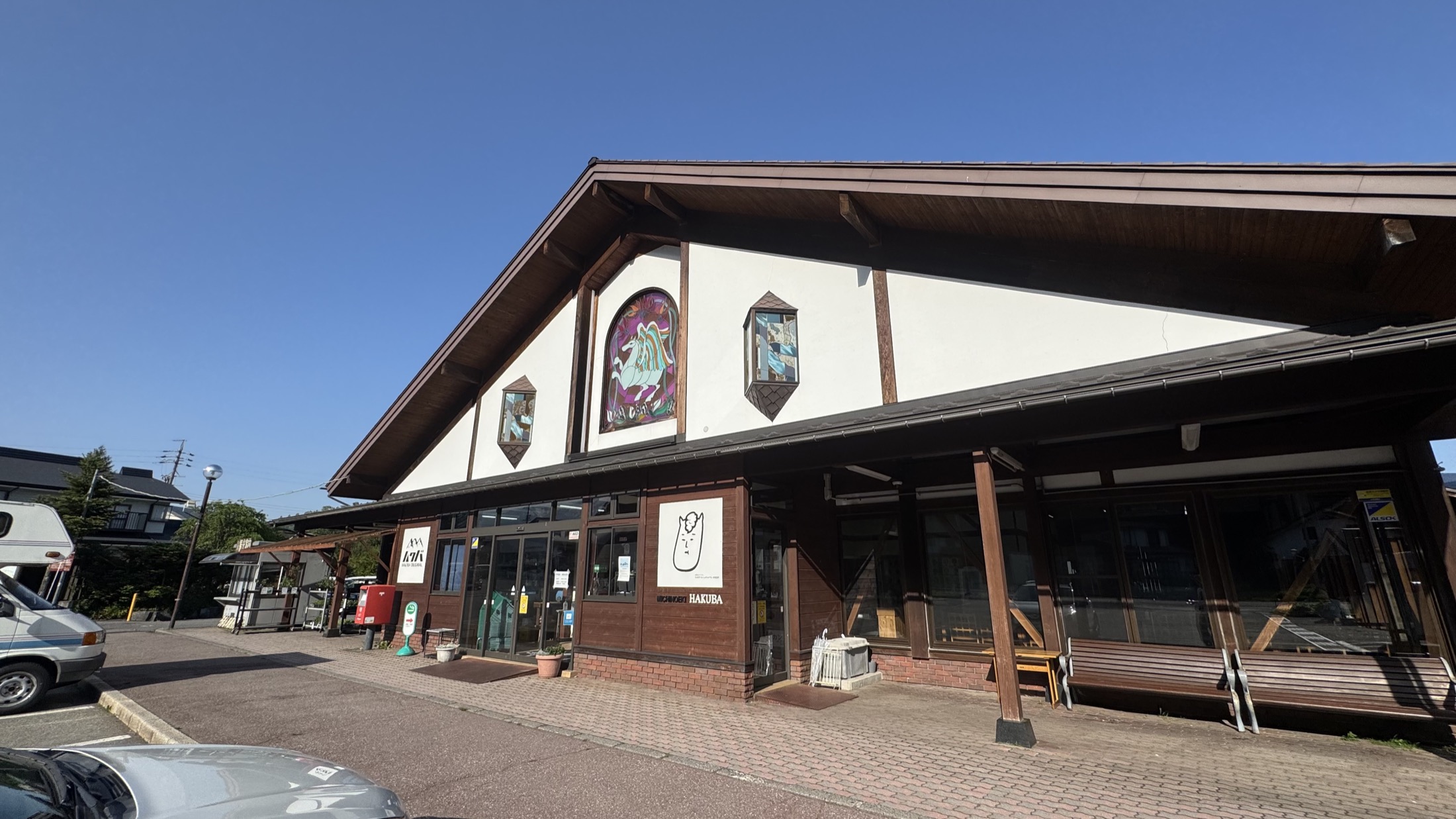 The chalet-style timber-and-plaster building of Michi-no-Eki Hakuba roadside station in Nagano under a clear blue sky, with stained-glass mountain artwork on the gable, a HAKUBA sign and a car park out front.