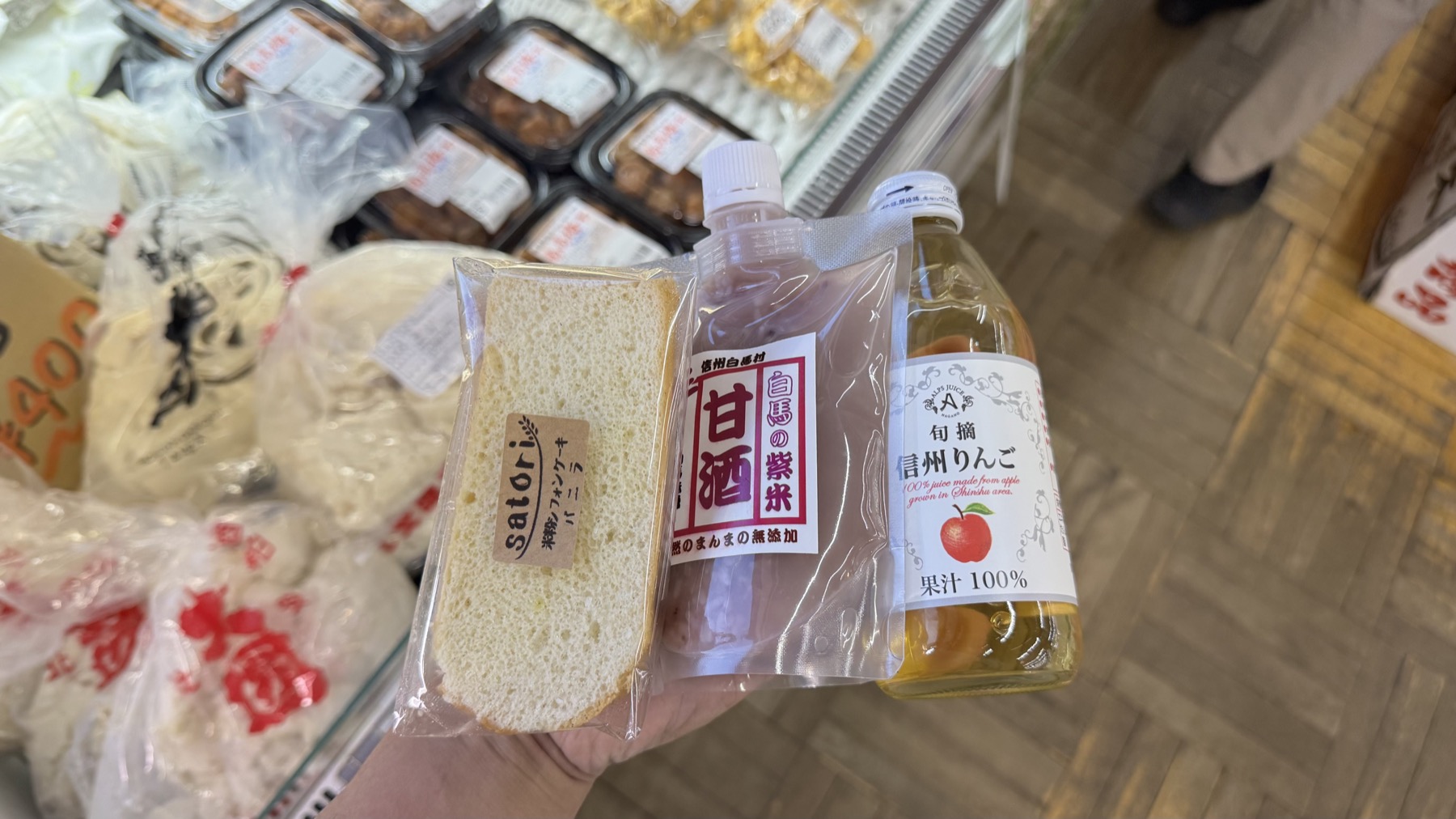 A hand holding three purchases from Michi-no-Eki Hakuba, a slice of rice-flour chiffon cake, a bottle of purple-rice amazake and a bottle of Shinshu apple juice, with packaged purple-rice onigiri on the shelf behind.