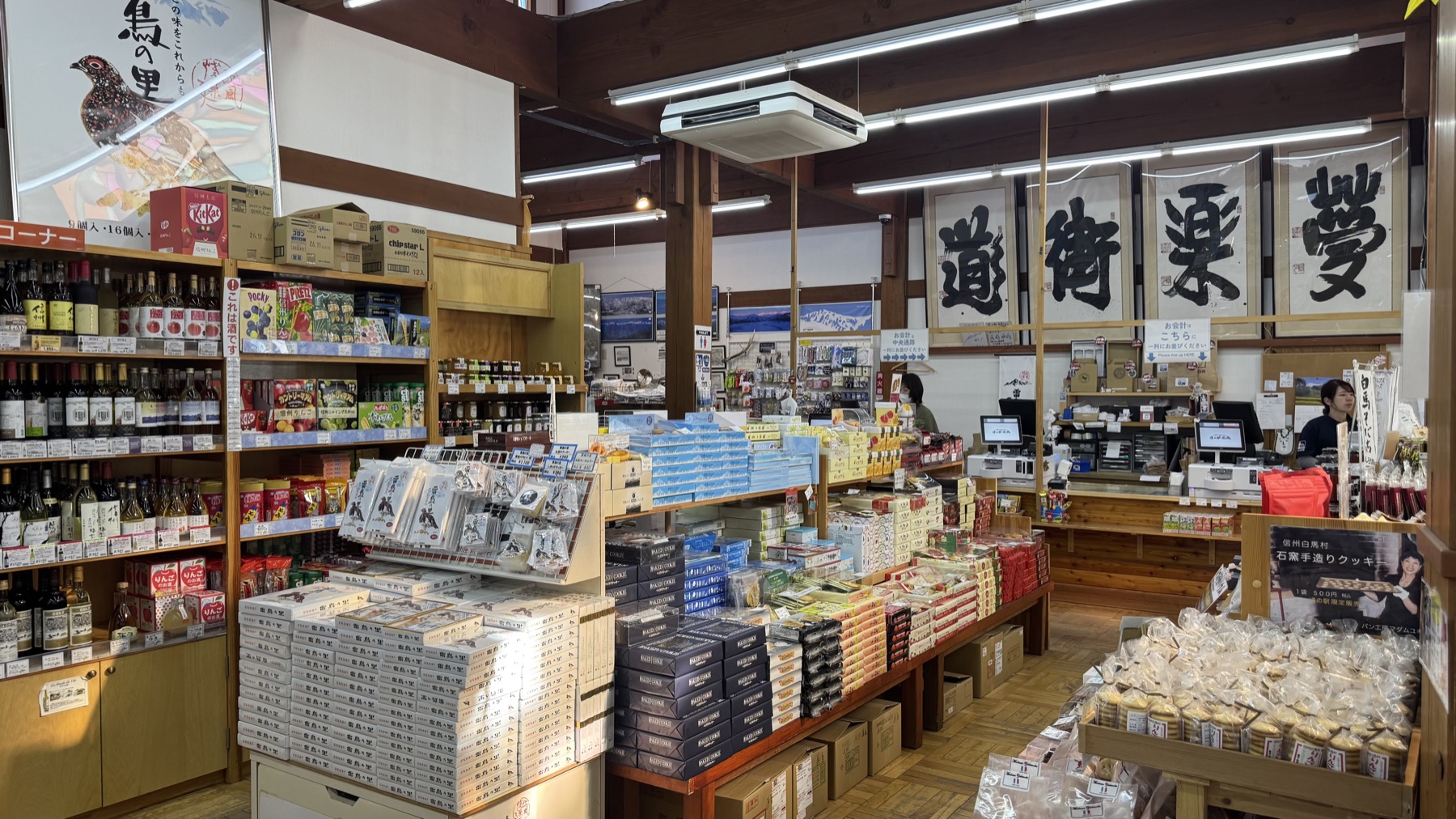 The interior of the Michi-no-Eki Hakuba farm shop, with wooden shelves of local bottles, boxed sweets and packaged goods, a staff counter on the right and large Japanese calligraphy banners on the wall.