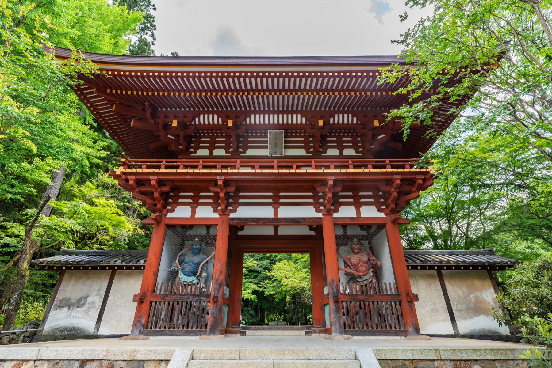 The two-story vermillion Niomon gate of Muro-ji with two carved Nio guardian statues set in its side bays, surrounded by fresh green maple leaves and seen from the stone steps below