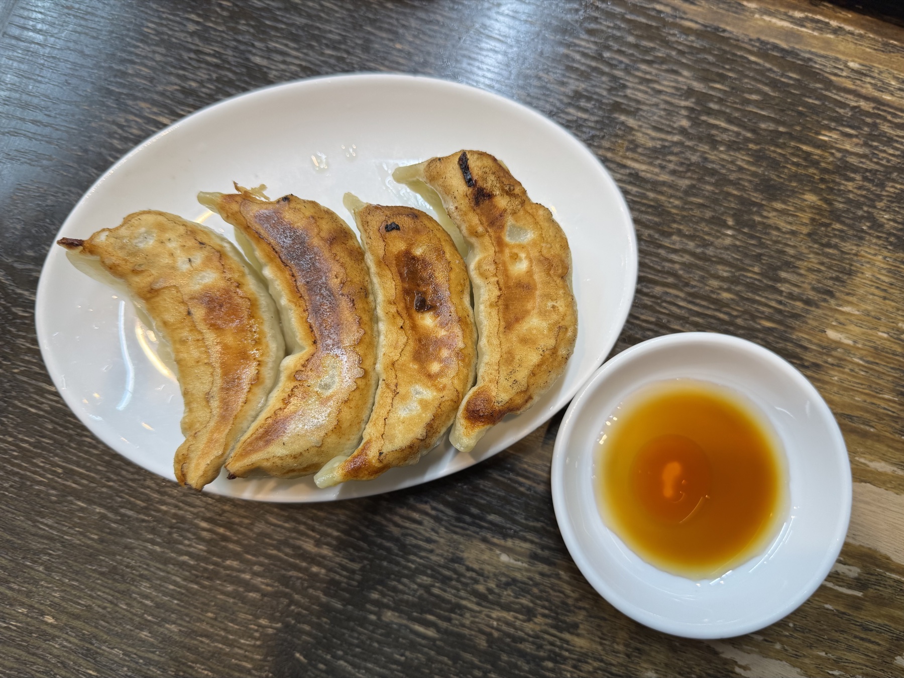 Four plump pan-fried jumbo gyoza dumplings on a white oval plate with a small dish of dipping sauce, the wrappers browned and blistered on the bottom.