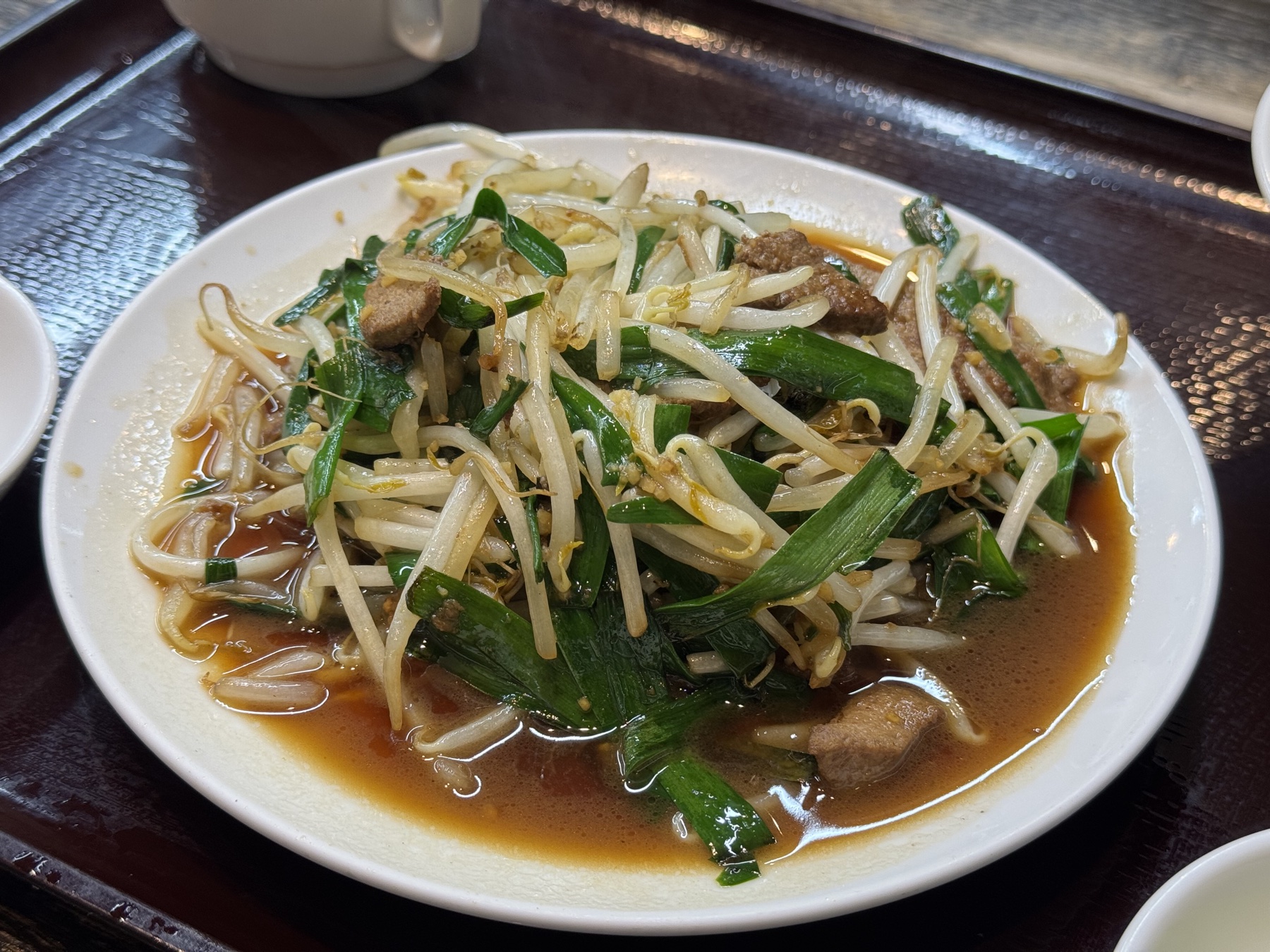 A saucy stir-fry of pork liver, bean sprouts and garlic chives piled on a white plate, glossy with a dark brown sauce, on a lacquered tray.