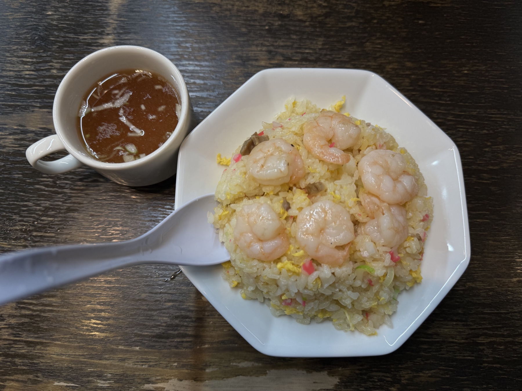 A domed plate of shrimp fried rice topped with several pink prawns and flecks of egg and kamaboko, served with a small mug of soup on a dark wooden table.