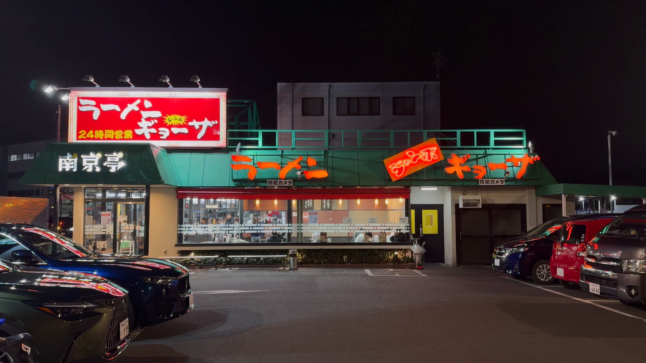 The Nankintei Kunitachi branch lit up at night, a green-roofed roadside Chinese diner with glowing red ramen and gyoza signs, a 24-hour notice and a wide car park out front in Kunitachi, Tokyo.